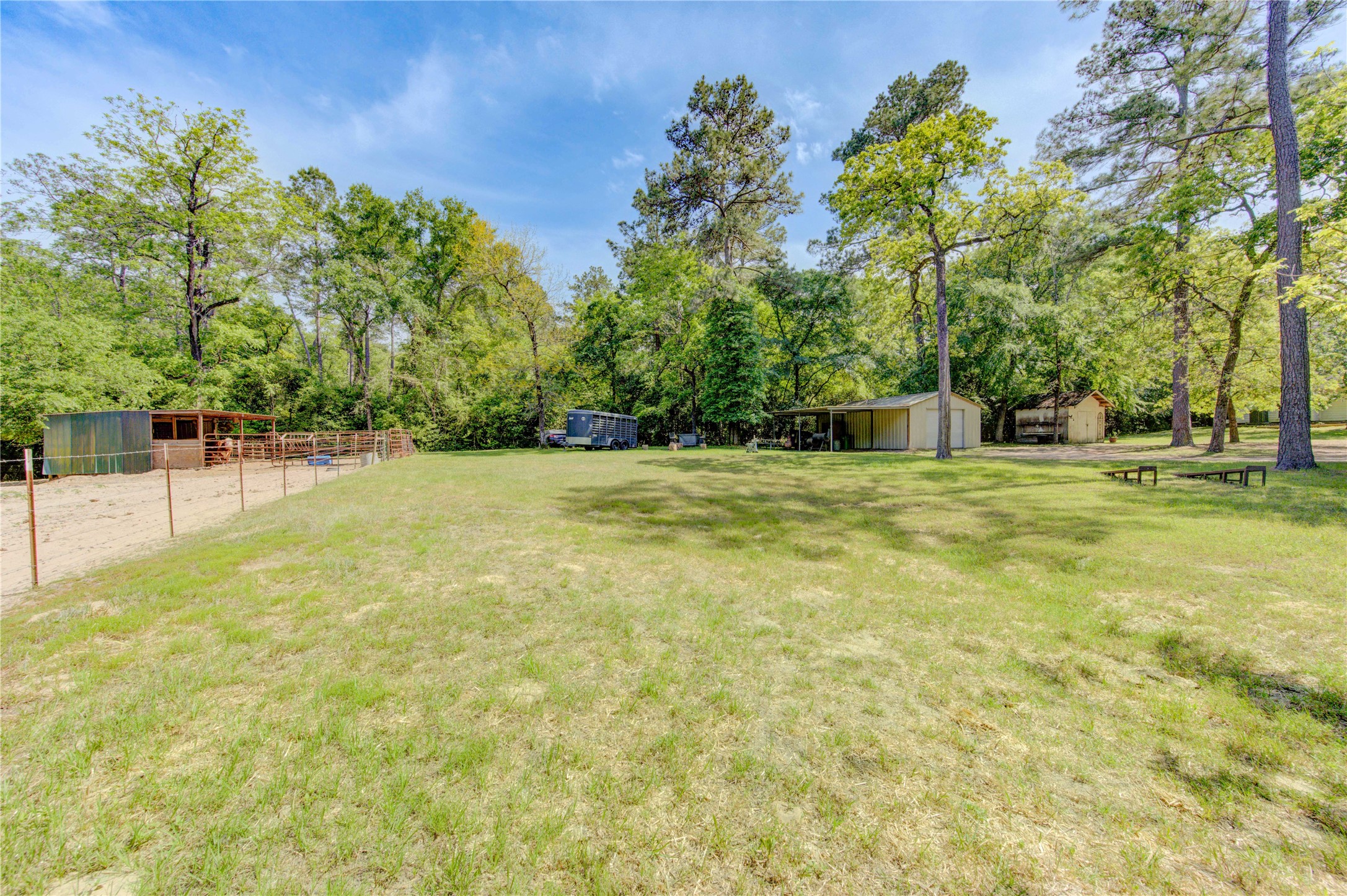 25903 Penguin Street Magnolia, TX 77355 - Photo 4 of 46 This spacious property features a large, open grassy area surrounded by mature trees, offering shade and privacy. There are multiple outbuildings, including a fenced area suitable for animals. Ideal for those seeking a blend of nature and utility.
