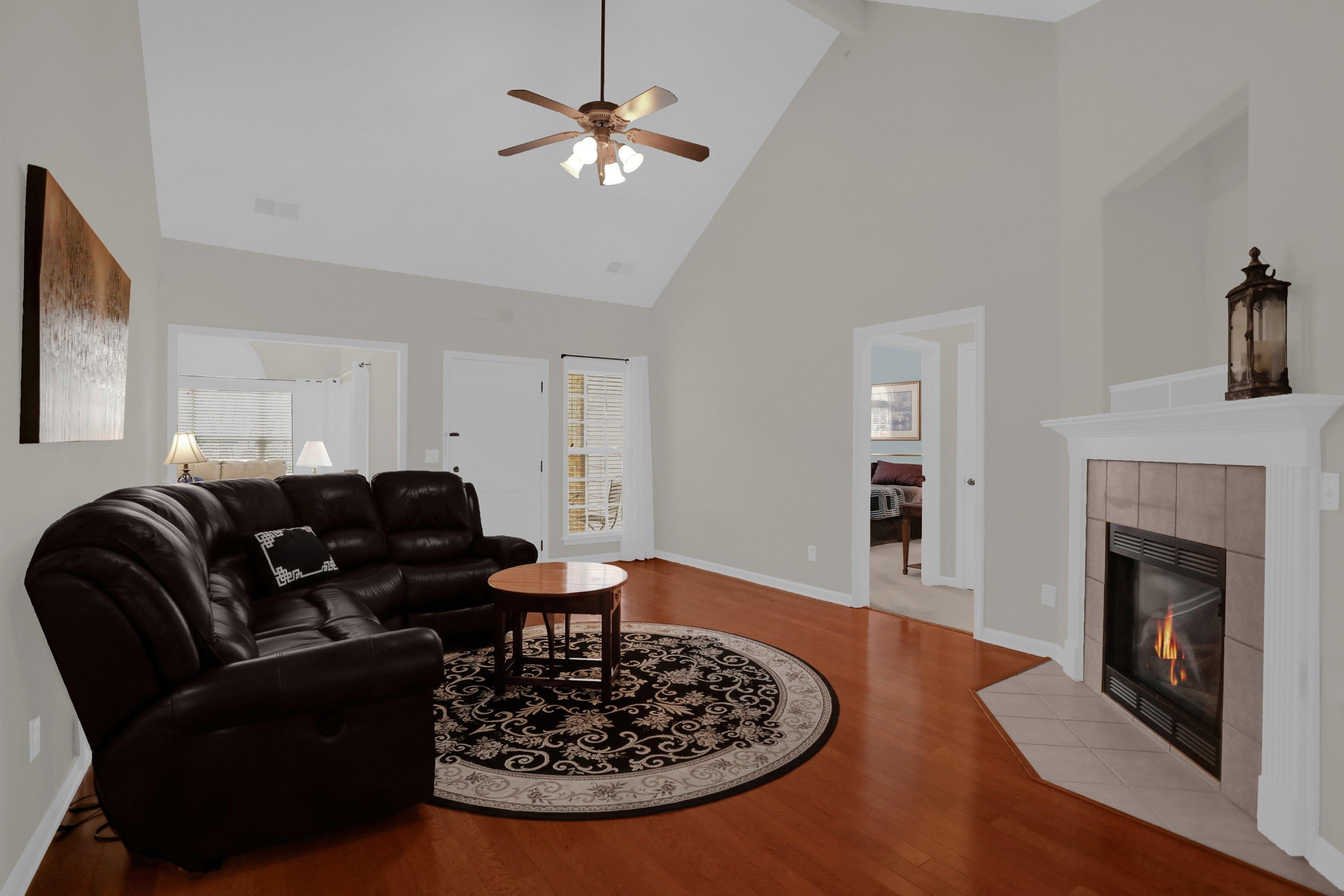 34 Peyton Ridge Drive, Unit 21 Collierville, TN 38017 - Photo 8 of 37 Living area featuring vaulted ceilings, wood-finish flooring, a ceiling fan with integrated lighting, a tiled fireplace with white mantel, and a doorway opening to an outdoor area