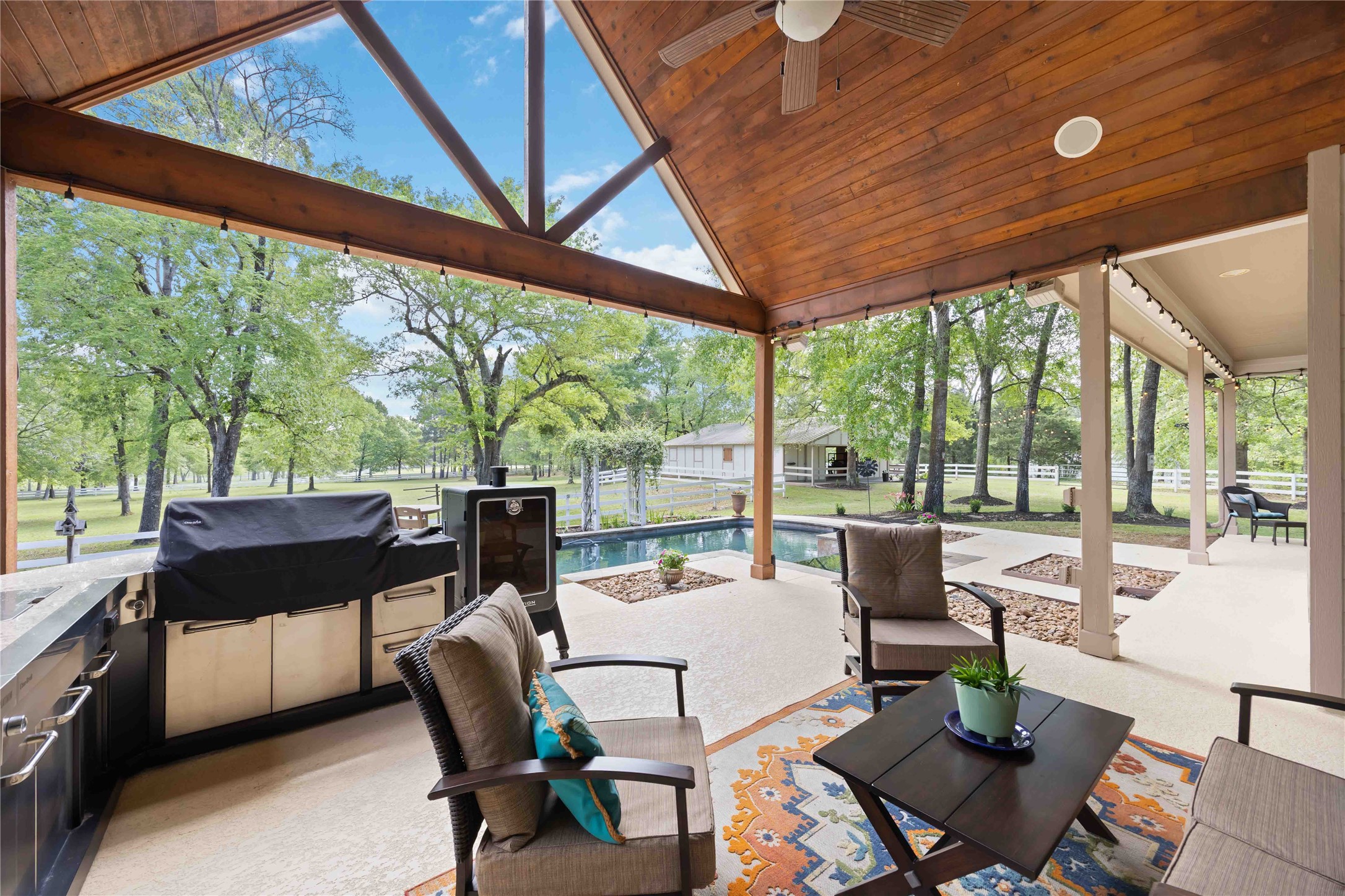 1809 St Beulah Chapel Road Montgomery, TX 77316 - Photo 36 of 50 This photo showcases a spacious, covered outdoor patio with comfortable seating, a built-in grill, and a ceiling fan. Overlooking a lush backyard with a pool and surrounded by mature trees, it offers a perfect space for relaxing and entertaining.