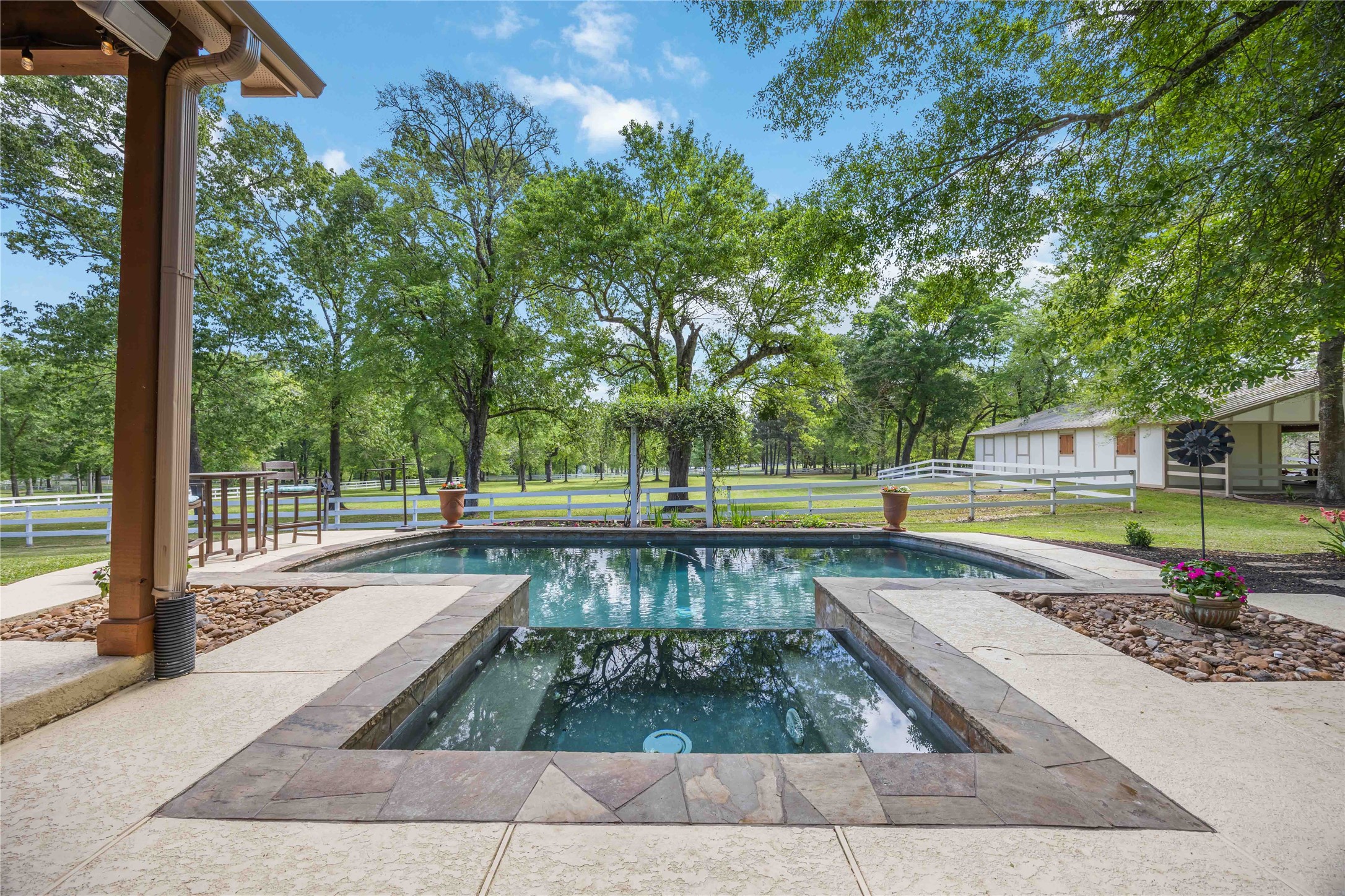 1809 St Beulah Chapel Road Montgomery, TX 77316 - Photo 40 of 50 This photo showcases a serene backyard with a spacious pool and spa, surrounded by lush greenery and mature trees. The view includes a well-maintained lawn, a white fence, and a barn-like structure, offering a picturesque, private retreat.