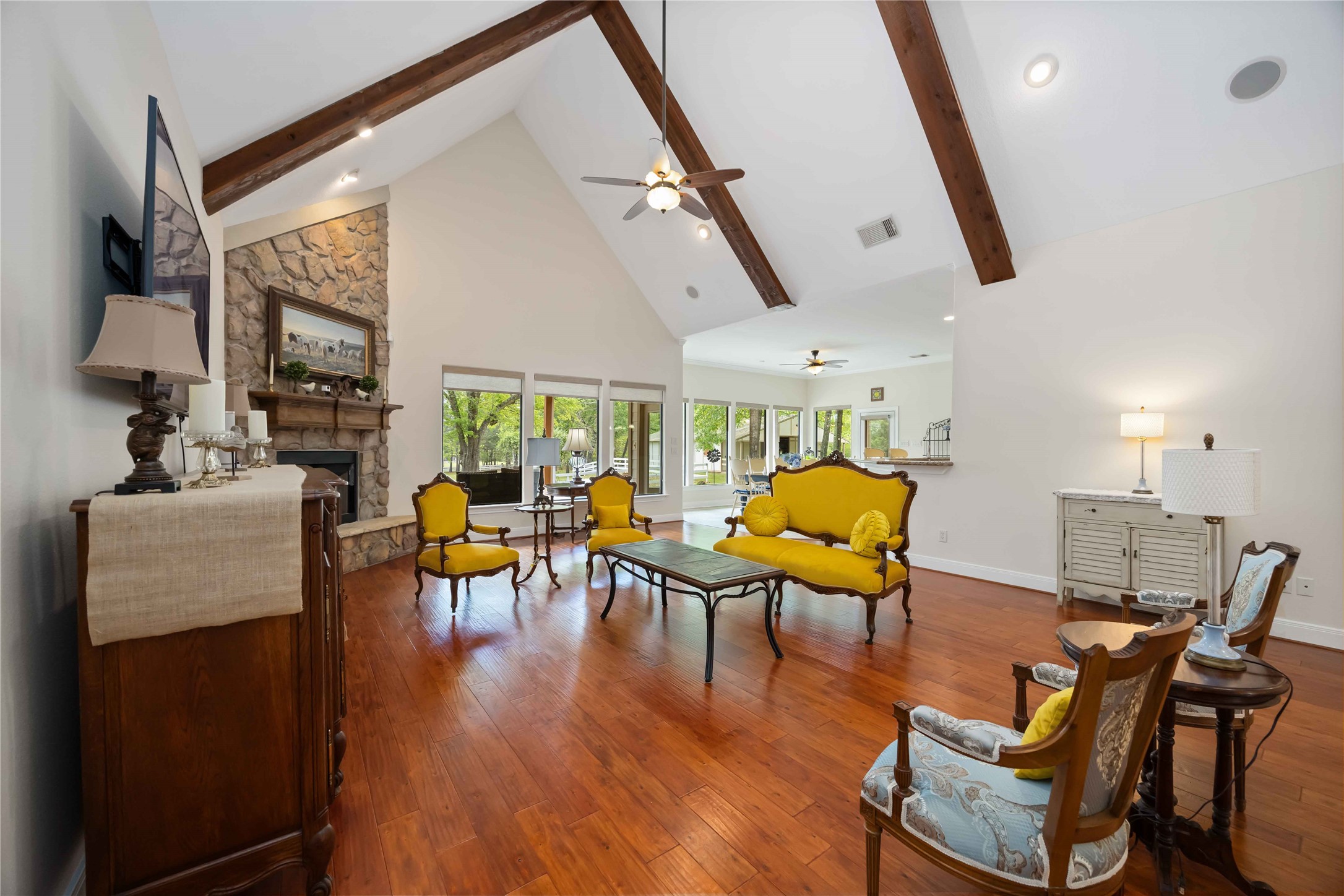 1809 St Beulah Chapel Road Montgomery, TX 77316 - Photo 9 of 50 This spacious living room features high vaulted ceilings with exposed wooden beams, a cozy stone fireplace, and large windows providing abundant natural light. The room is tastefully furnished with elegant seating and hardwood floors, creating a warm and inviting atmosphere.