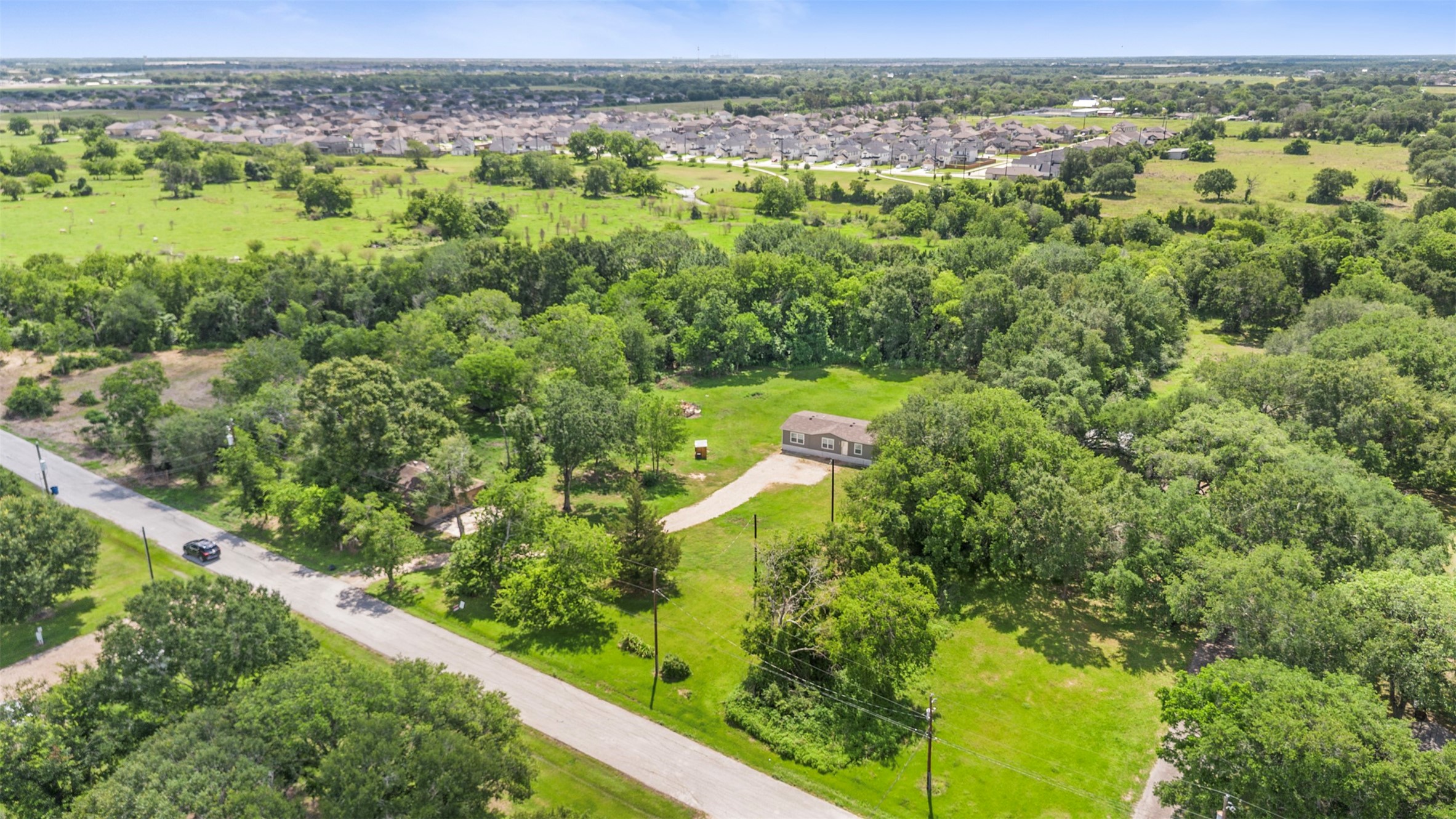 2701 Mazen Road Rosenberg, TX 77471 - Photo 9 of 10 This aerial view shows a spacious rural property surrounded by lush greenery and trees. A single house is situated on a large plot, providing privacy. A road runs alongside the property, with a suburban neighborhood visible in the distance.