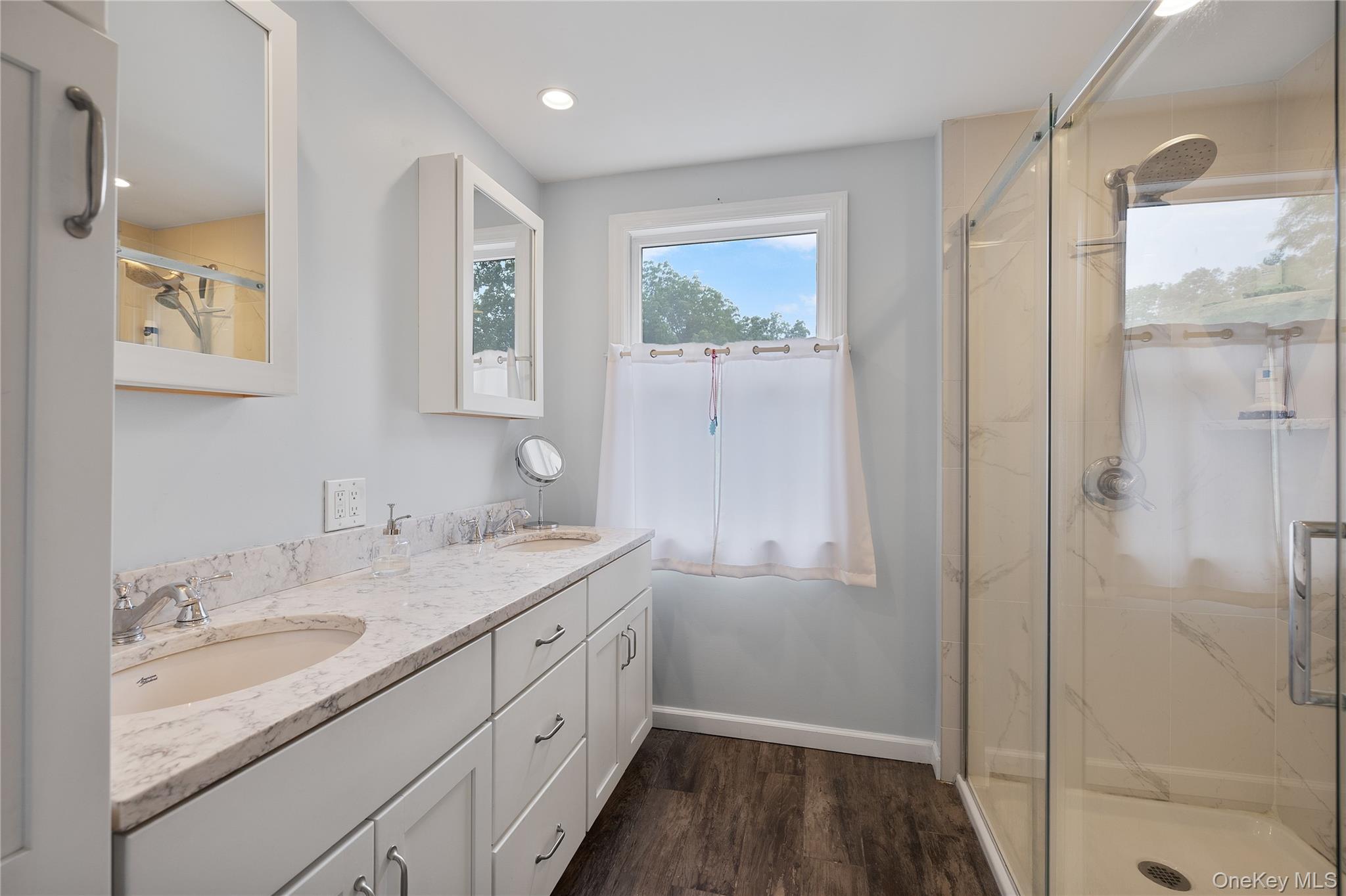 9 Three Sisters Road St. James, NY 11780 - Photo 13 of 36 Primary updated bathroom featuring a dual vanity with white cabinetry and light-toned countertops, a glass-enclosed shower with a rainfall showerhead, and wood-finish flooring