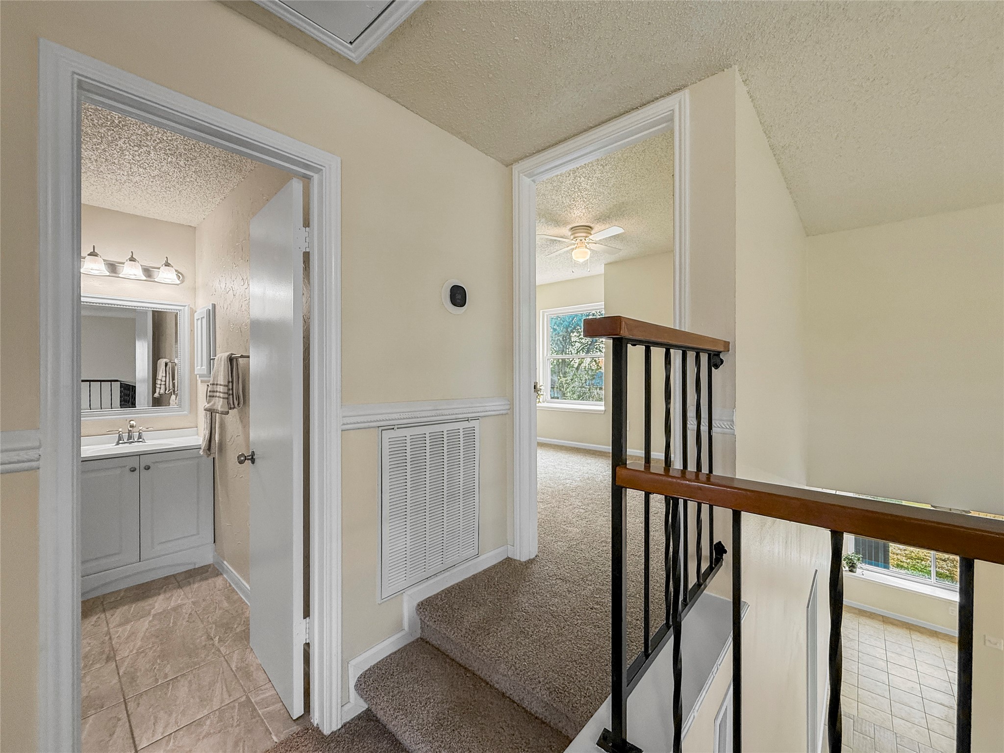 13635 Repa Lane Houston, TX 77014 - Photo 41 of 49 This photo shows a small landing area with carpeted stairs leading to a bathroom and a bedroom. The bathroom features tile flooring, a vanity with a mirror, and three overhead lights. The bedroom has a window letting in natural light and a ceiling fan. The area is well-lit and has neutral-colored walls.