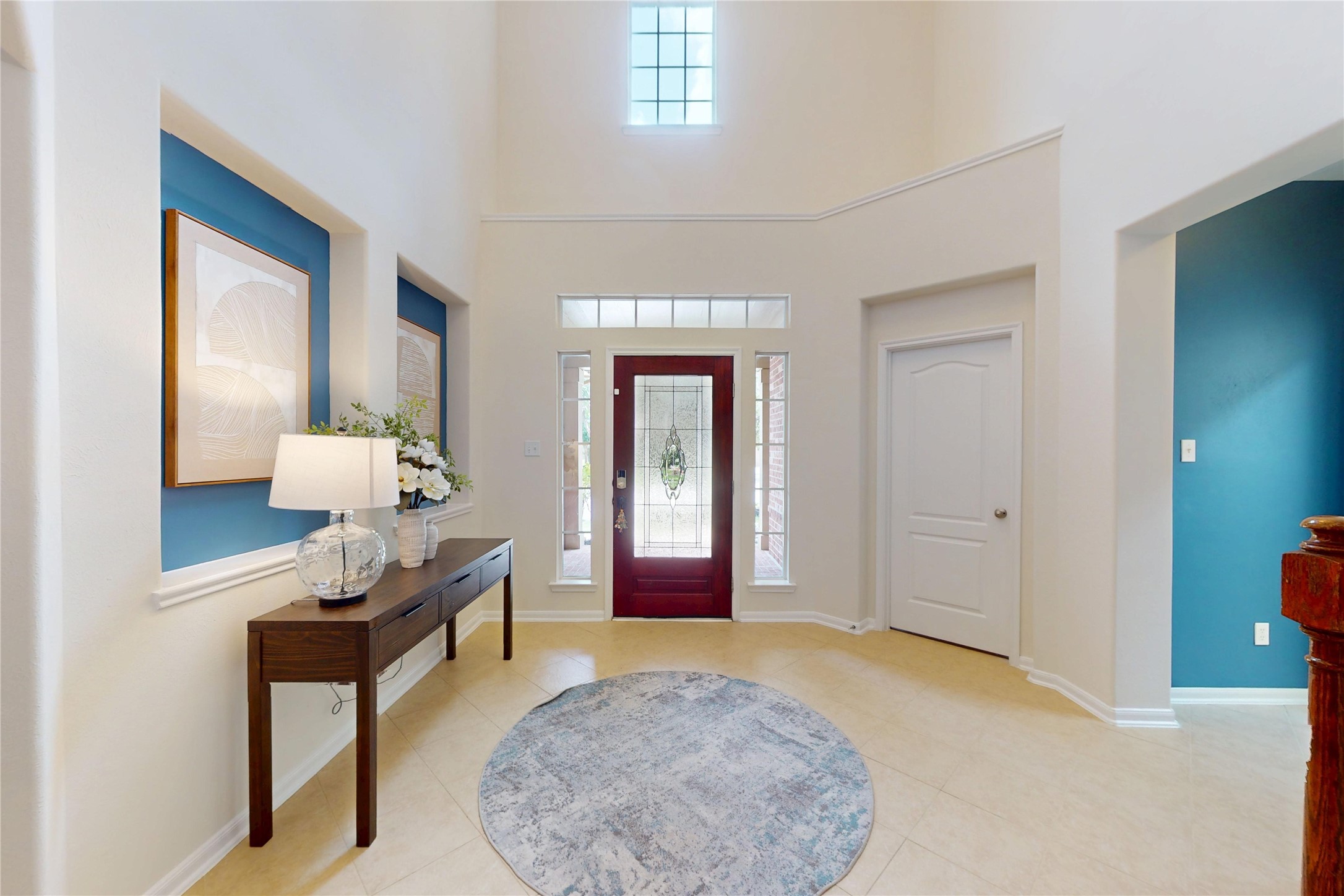 17402 Rainer Valley Lane Humble, TX 77346 - Photo 2 of 33 This entryway features high ceilings and abundant natural light from a large window above. A stylish front door with glass accents is flanked by sidelight windows. There’s a simple, elegant console table with decorative pieces, and the walls have modern art and a contrasting blue accent. The space feels open and welcoming.