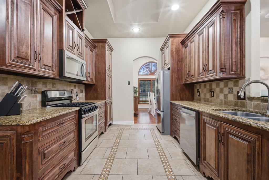 603 Cedar Drive Pilot Point, TX 76258 - Photo 6 of 39 Galley kitchen featuring rich wood cabinetry, stainless steel appliances, speckled stone countertops, and a decorative tile backsplash