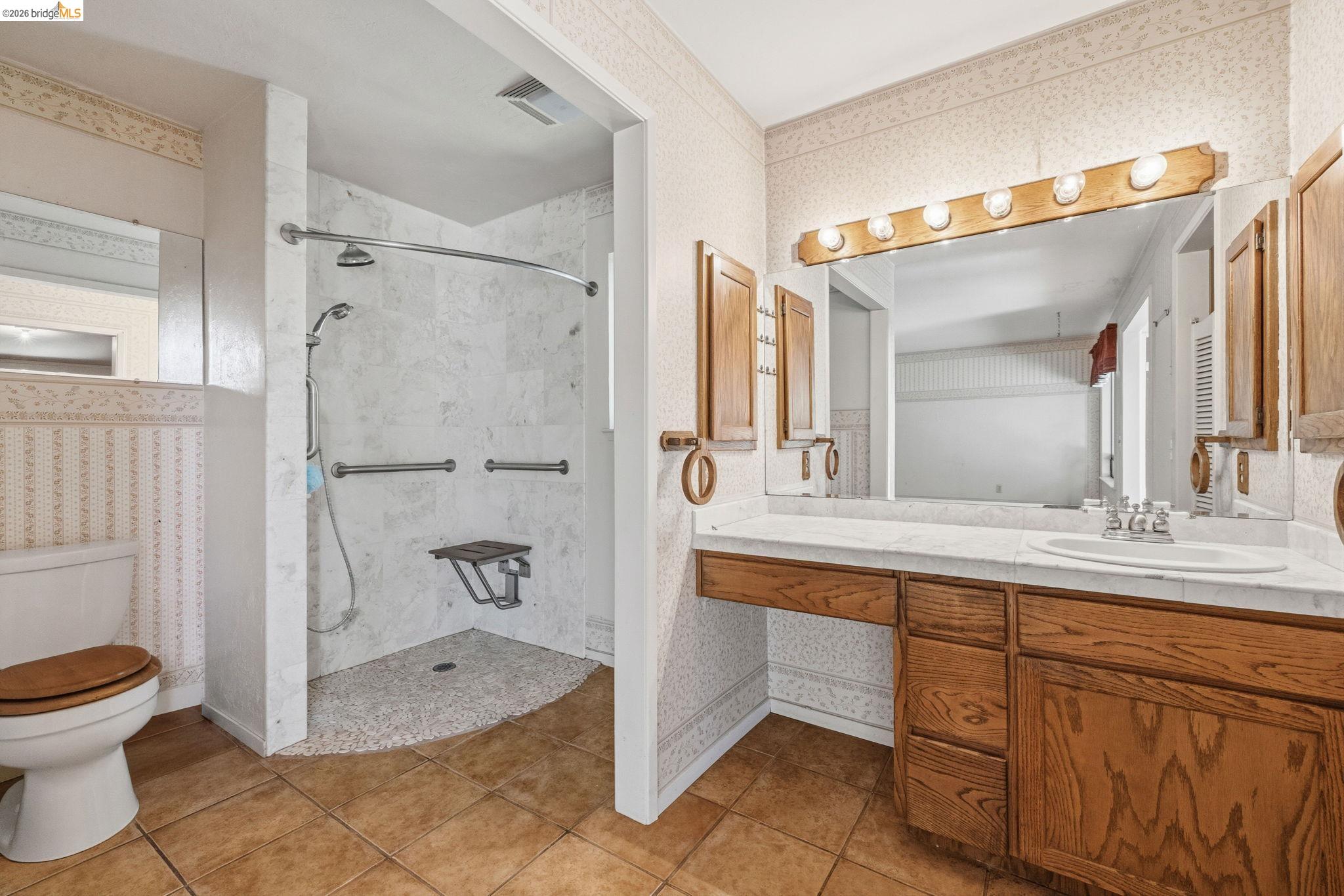 22401 South Fork Road Sonora, CA 95370 - Photo 26 of 34 Bathroom featuring a spacious walk-in shower with grab bars and a mounted seat, a large vanity with an integrated sink, ample wood cabinetry, a generous wall mirror, and vanity lighting