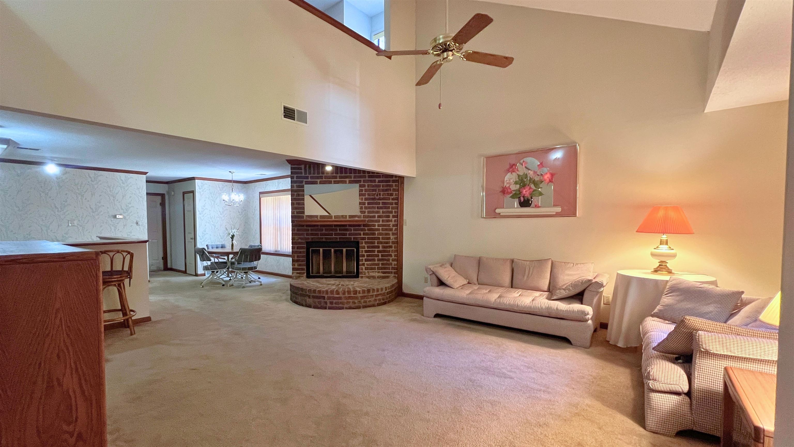 Living room featuring brick wall, a fireplace, a towering ceiling, carpet flooring, and ceiling fan