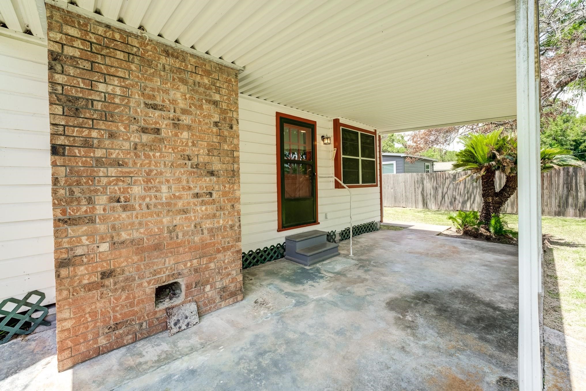 305 Yaupon Street Sweeny, TX 77480 - Photo 2 of 22 This photo showcases a covered patio area featuring a brick wall and a small step leading to a back door. The space opens to a fenced backyard with greenery, providing a private and sheltered outdoor area.
