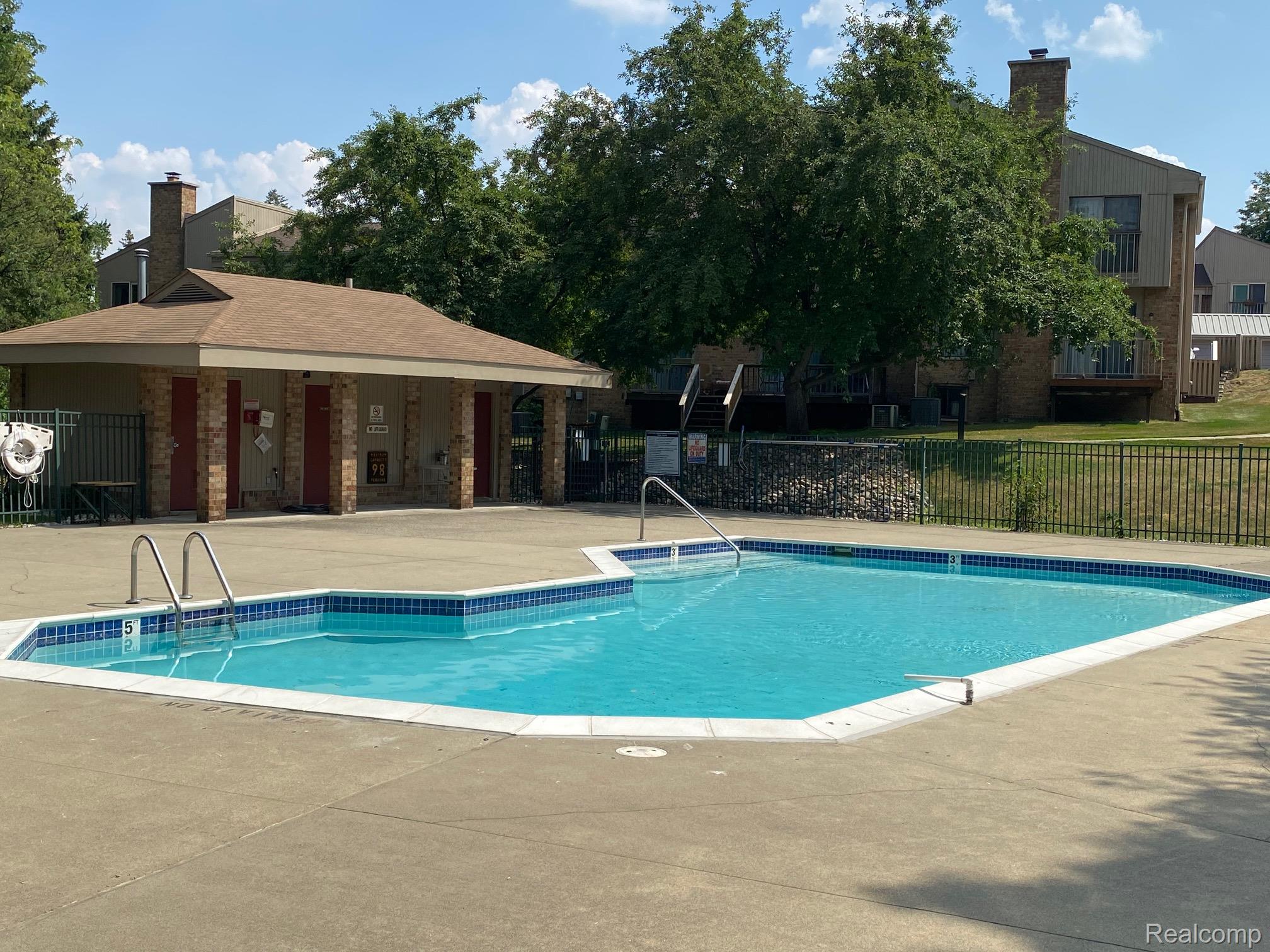 629 Oakbrook West, Unit 18 Rochester, MI 48307 - Photo 6 of 18 Community swimming pool with concrete decking, a surrounding metal fence, and a nearby brick-clad utility building