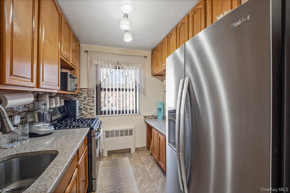 155-14 86th Street, Unit 2 Queens, NY 11414 - Photo 7 of 14 Kitchen with stainless steel appliances, light stone counters, brown cabinetry, radiator heating unit, and backsplash