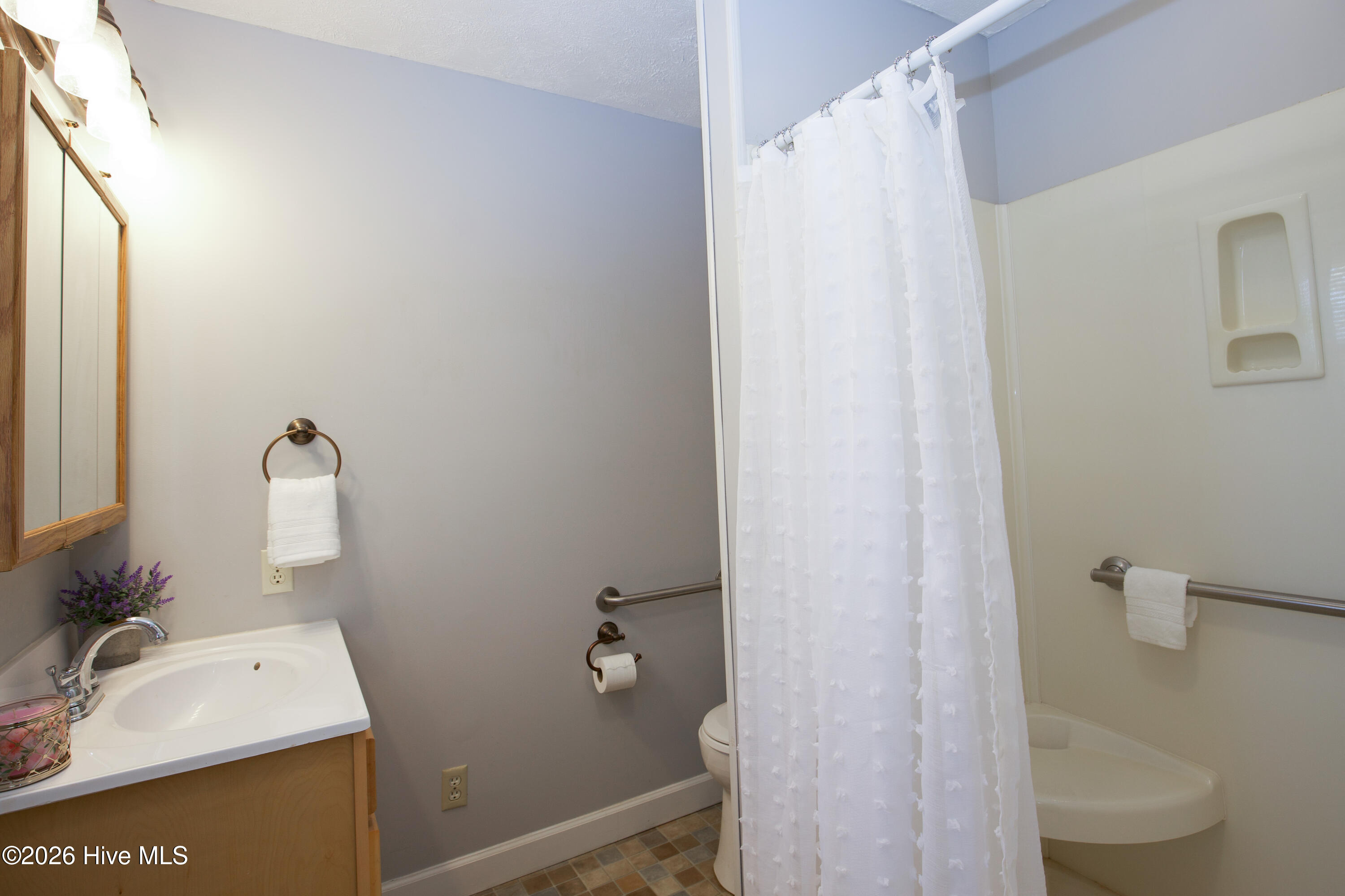 5068 Lobelia Road Vass, NC 28394 - Photo 23 of 70 The primary bathroom offers a convenient walk-in shower with seating and safety grab bars, ideal for accessibility and comfort. A crisp white vanity with mirrored medicine cabinet and bronze fixtures complements the space. Neutral tile flooring adds warmth and durability.