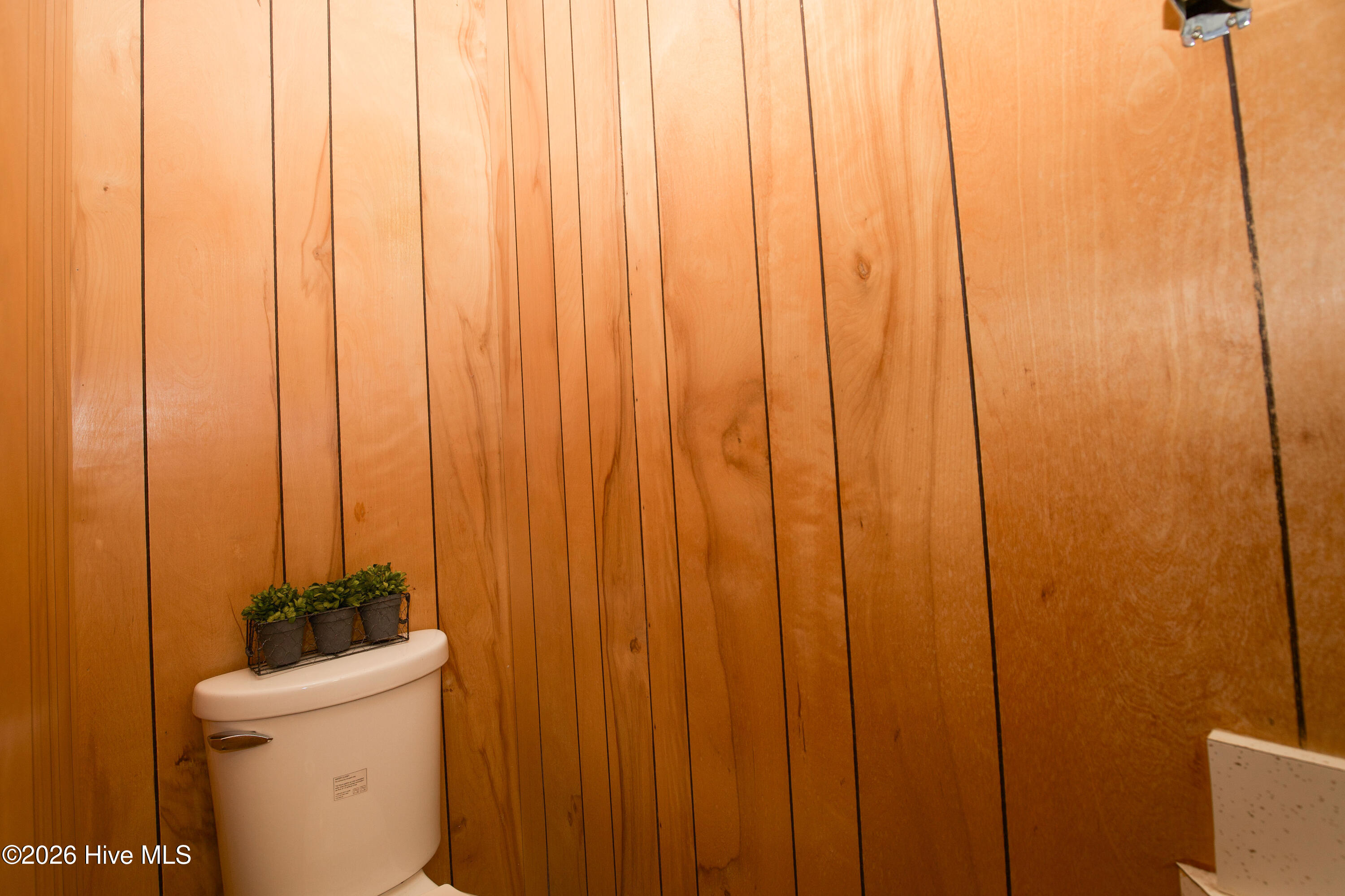 5068 Lobelia Road Vass, NC 28394 - Photo 53 of 70 This photo shows the private water closet area located within the spacious laundry and utility room. It features a standard toilet set against full-height retro wood paneling, blending functionality with a nostalgic aesthetic. The vertical wood grain creates visual warmth and adds a touch of vintage charm to this practical corner of the home. A decorative plant accent on the toilet tank adds a cozy, lived-in feel. This tucked-away bathroom area is convenient for multitasking while doing laundry or working around the house.