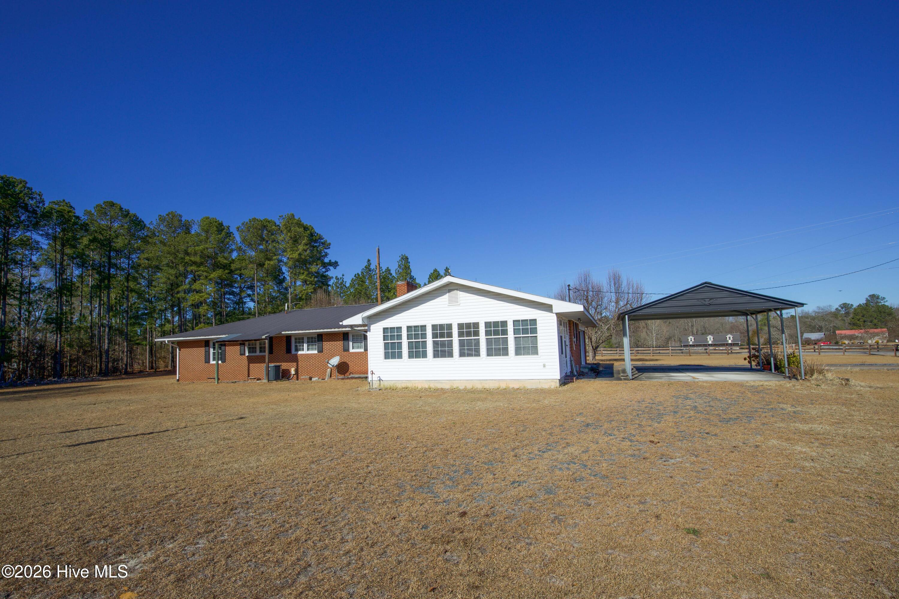 5068 Lobelia Road Vass, NC 28394 - Photo 57 of 70 Rear view of 5068 Lobelia Rd showing the white Carolina Room, metal-roofed carport, and fenced backyard area. The Carolina Room, featuring abundant natural light, extends from the main brick structure, offering extra flexible living space. The carport provides covered parking and convenience, with easy access into the side entrance of the home. The open yard space surrounding the back of the property offers plenty of options for future garden beds, recreational use, or outdoor gatherings. Surrounded by mature trees, the backyard blends privacy with functionality.