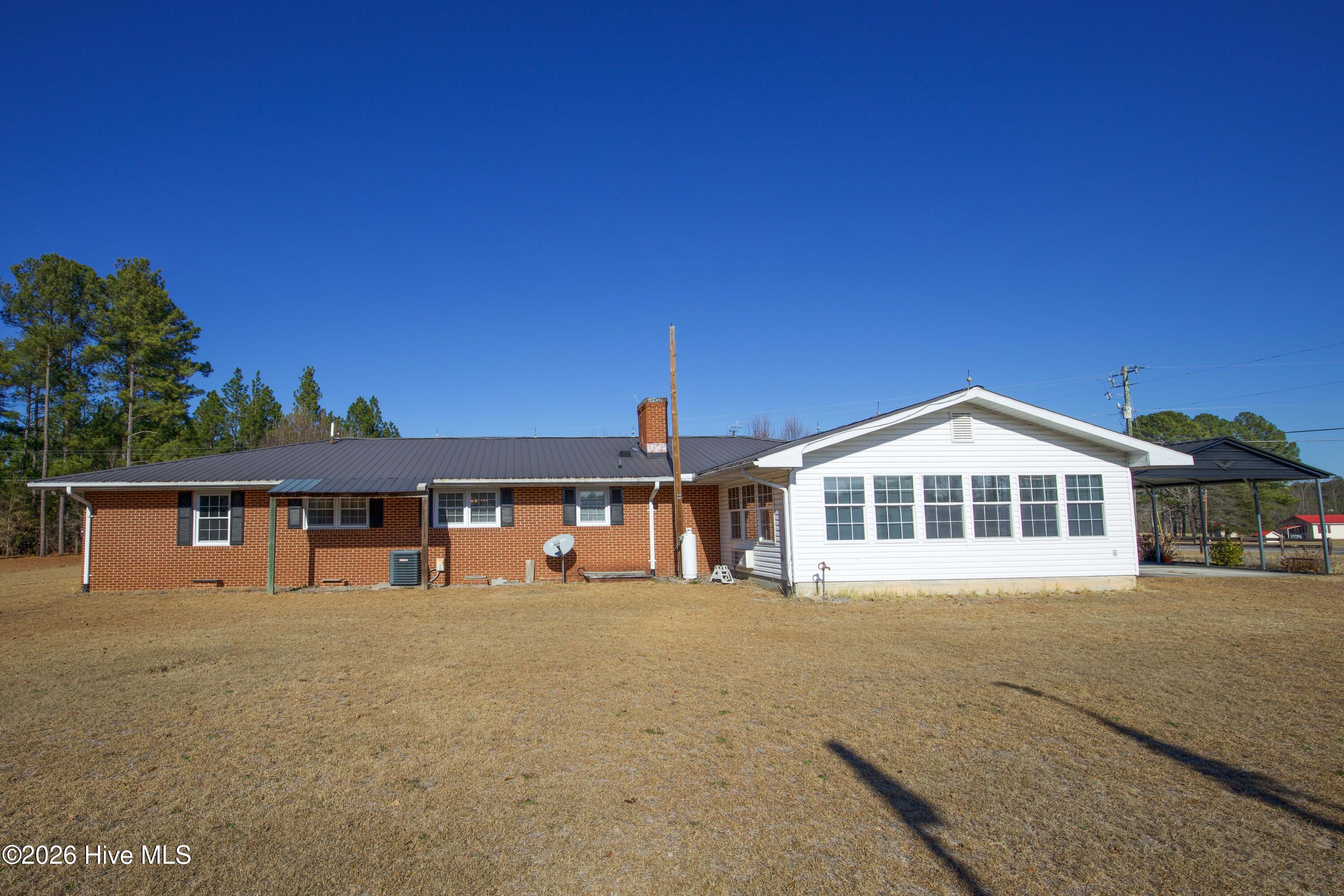 5068 Lobelia Road Vass, NC 28394 - Photo 58 of 70 This rear-facing photo highlights the full exterior of the Carolina Room addition and the back side of 5068 Lobelia Rd. The contrast between the brick structure and white siding offers a clear view of the home’s layout and expanded footprint. You’ll see the metal roof spanning the main home, dual HVAC units, propane tank, and satellite dish setup. The Carolina Room’s large windows face out over the backyard, letting in abundant light and offering clear views of the spacious lot. Also visible is the entry to the covered parking area.