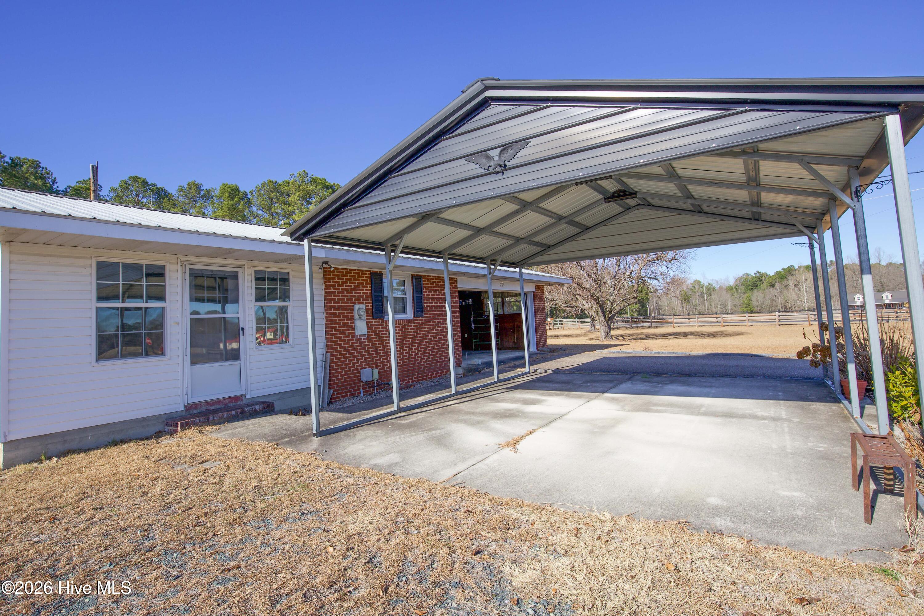 5068 Lobelia Road Vass, NC 28394 - Photo 59 of 70 This image captures the metal-framed covered parking area at the rear of 5068 Lobelia Rd, ideal for vehicle protection from the elements. The concrete pad extends into a driveway area, and the covered space provides convenient access to both the brick side entry and the Carolina Room entrance. The surrounding open land and fencing further highlight the spacious rural setting. This area supports practical use while blending into the home’s efficient footprint.