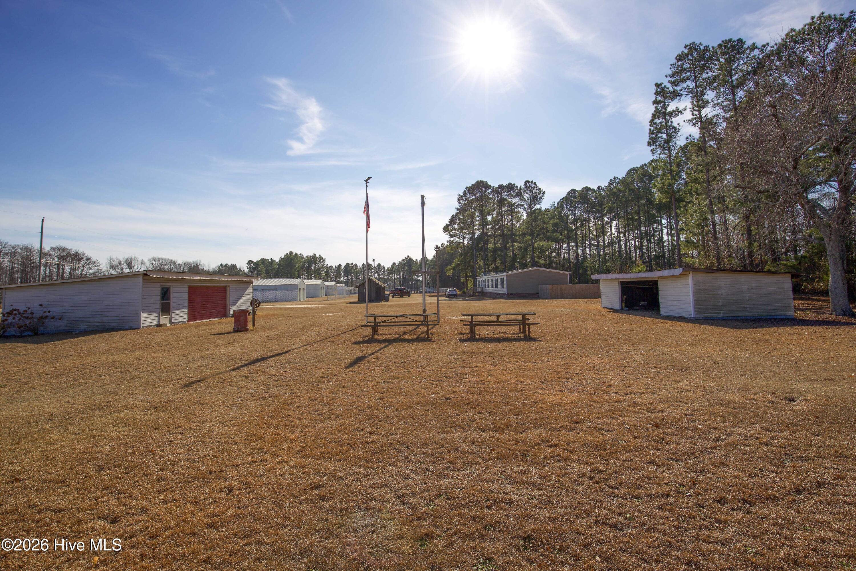5068 Lobelia Road Vass, NC 28394 - Photo 63 of 70 Expansive backyard view at 5068 Lobelia Rd featuring multiple outbuildings ideal for workshop space, storage, or equipment use. Two picnic tables are centrally placed near dual flagpoles, creating an open recreational area perfect for gatherings. Structures to the left and right offer flexibility for tools, hobby use, or agricultural needs. Bordered by tall pines and neighboring structures, this large lot delivers versatility and open sky views in a peaceful rural setting.