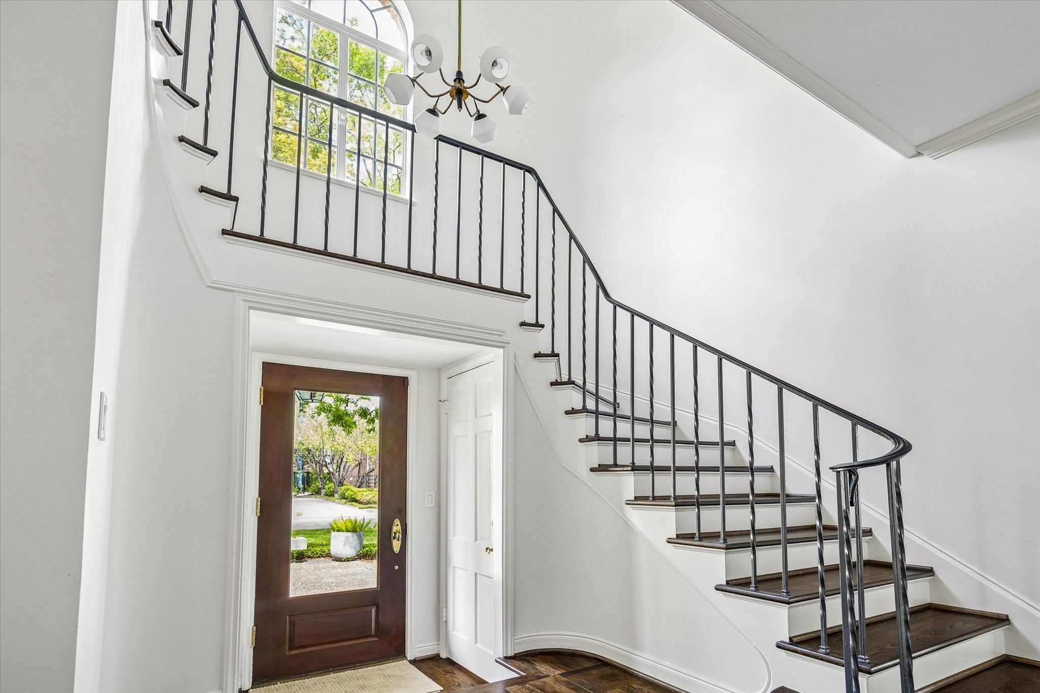 2133 Inwood Drive Houston, TX 77019 - Photo 3 of 22 Bright and inviting entryway with a curved staircase and elegant railing, large window, and modern chandelier. The wooden front door features a glass panel, offering a view of the greenery outside.