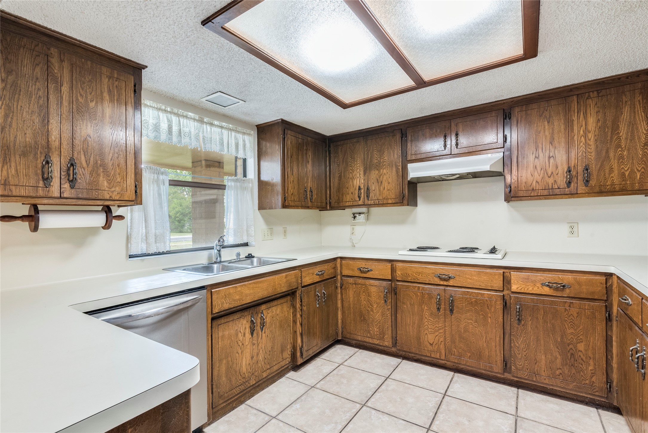 13505 Broadmeade Avenue Austin, TX 78729 - Photo 7 of 21 Kitchen with a textured ceiling, light countertops, stainless steel dishwasher, light tile patterned floors, and wood finish cabinets