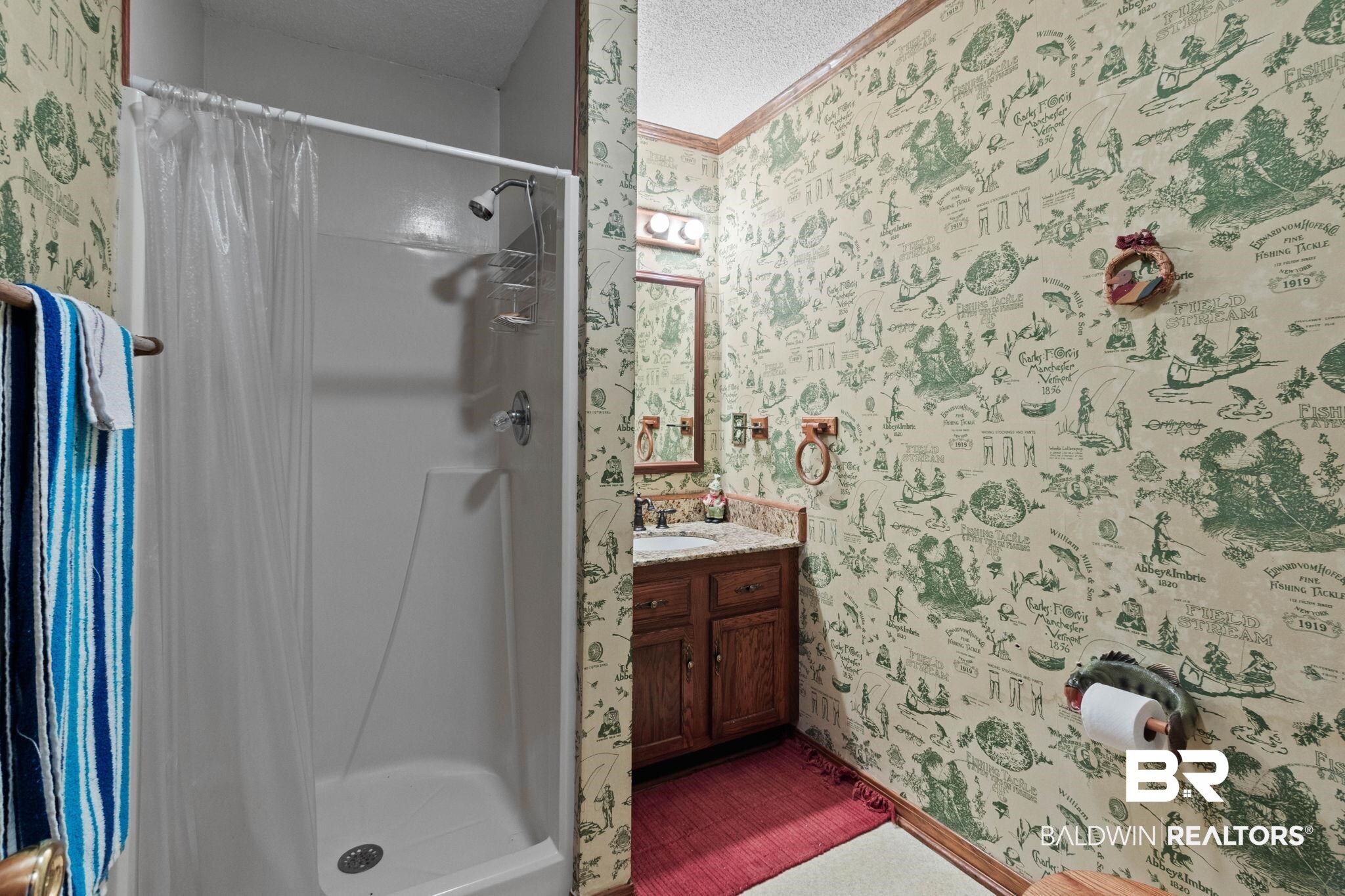 8153 Bryant's Lndg Road Stockton, AL 36579 - Photo 47 of 87 Bathroom featuring a white shower enclosure with chrome fixtures, a wood-finish vanity with a light-toned countertop, a framed mirror, and decorative wallpaper