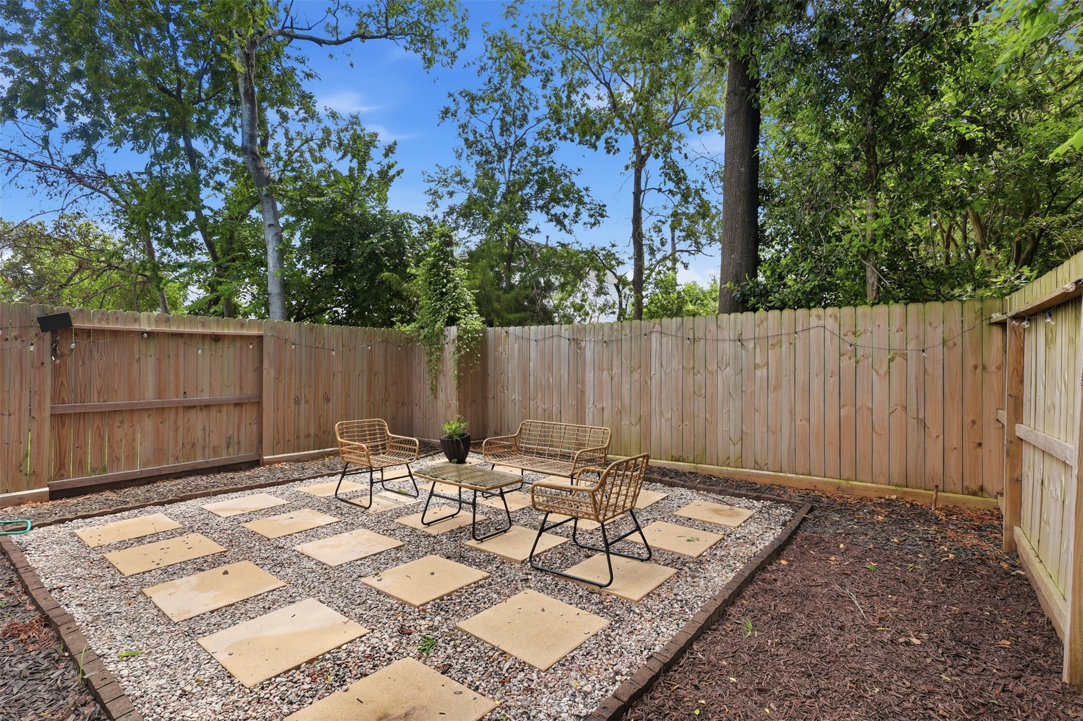 6833 Paris Street Houston, TX 77021 - Photo 22 of 23 This photo showcases a charming backyard patio area with a wooden privacy fence, surrounded by lush trees. The space features a grid of stone pavers set in gravel, with outdoor seating and a small table, creating an inviting spot for relaxation.