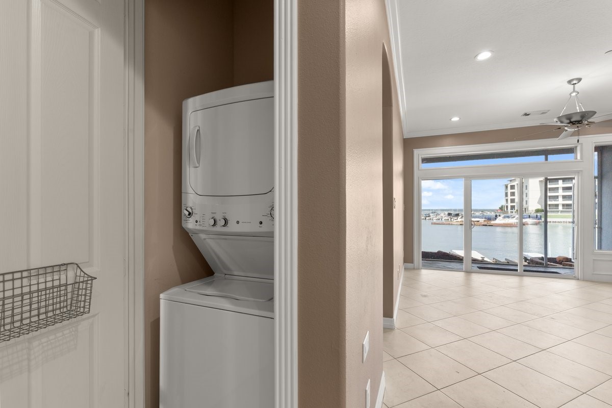 168 Lake Point Boulevard, Unit C102 Conroe, TX 77356 - Photo 24 of 35 This photo shows a well-lit living space with large windows offering a water view. It features a tile floor and a ceiling fan. A compact laundry area with a stacked washer and dryer is tucked into a closet.