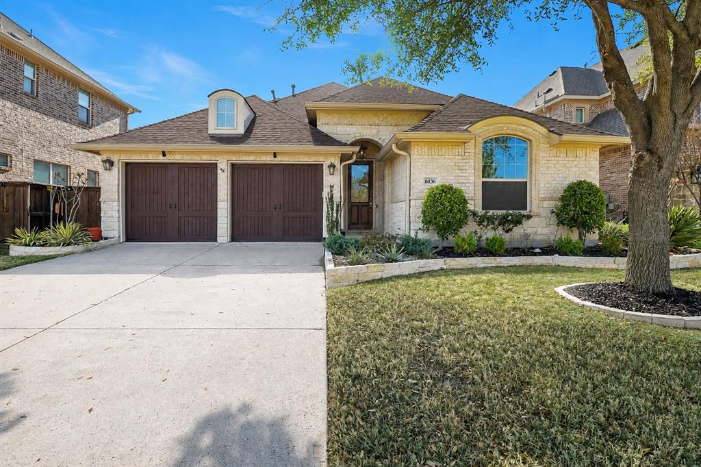 8036 Sylvan Dale Road Frisco, TX 75036 - Photo 1 of 1 Light-colored brick exterior with stone accents, two-car garage with dark wood-finish doors, arched window, and a prominent dormer
