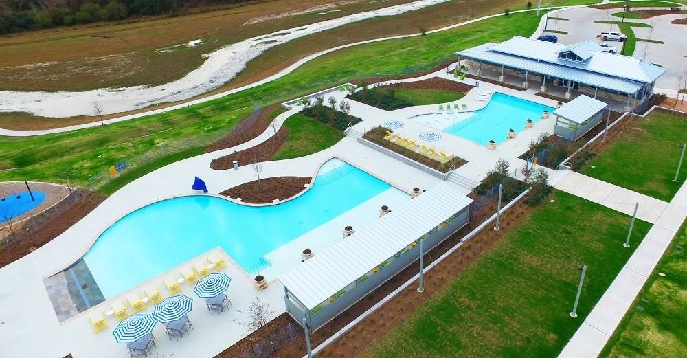 4806 Cedar Butte Lane Manvel, TX 77578 - Photo 18 of 18 This photo showcases a community recreation area featuring two large pools with ample seating, umbrellas, and a modern clubhouse. The surrounding landscape includes well-maintained green spaces and a parking area, offering a serene and inviting environment.