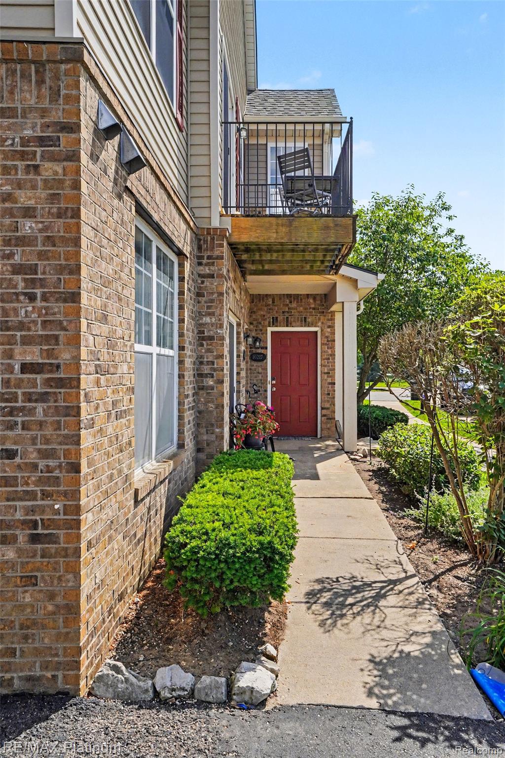10217 Crossview Trail Howell, MI 48855 - Photo 4 of 26 Brick and siding exterior featuring a red entry door, an upper-level balcony with black metal railing, a concrete walkway, and established landscaping