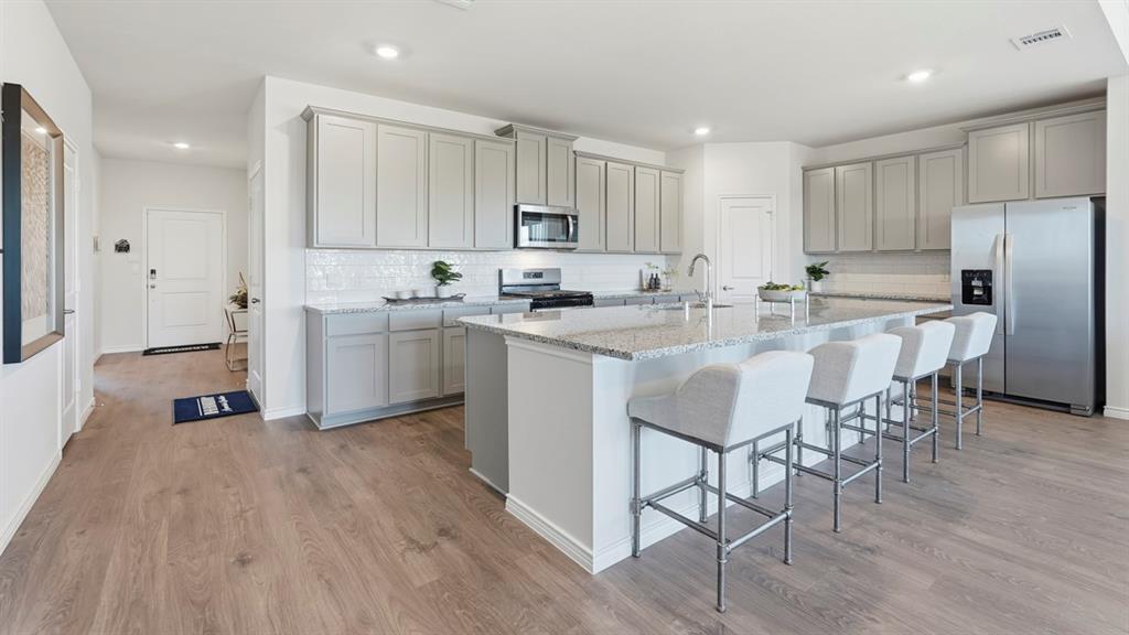 4197 Waldron Trail Forney, TX 75126 - Photo 2 of 25 Contemporary kitchen featuring grey cabinetry, stainless steel appliances, a subway tile backsplash, a large center island with a light-toned countertop, and wood-finish flooring