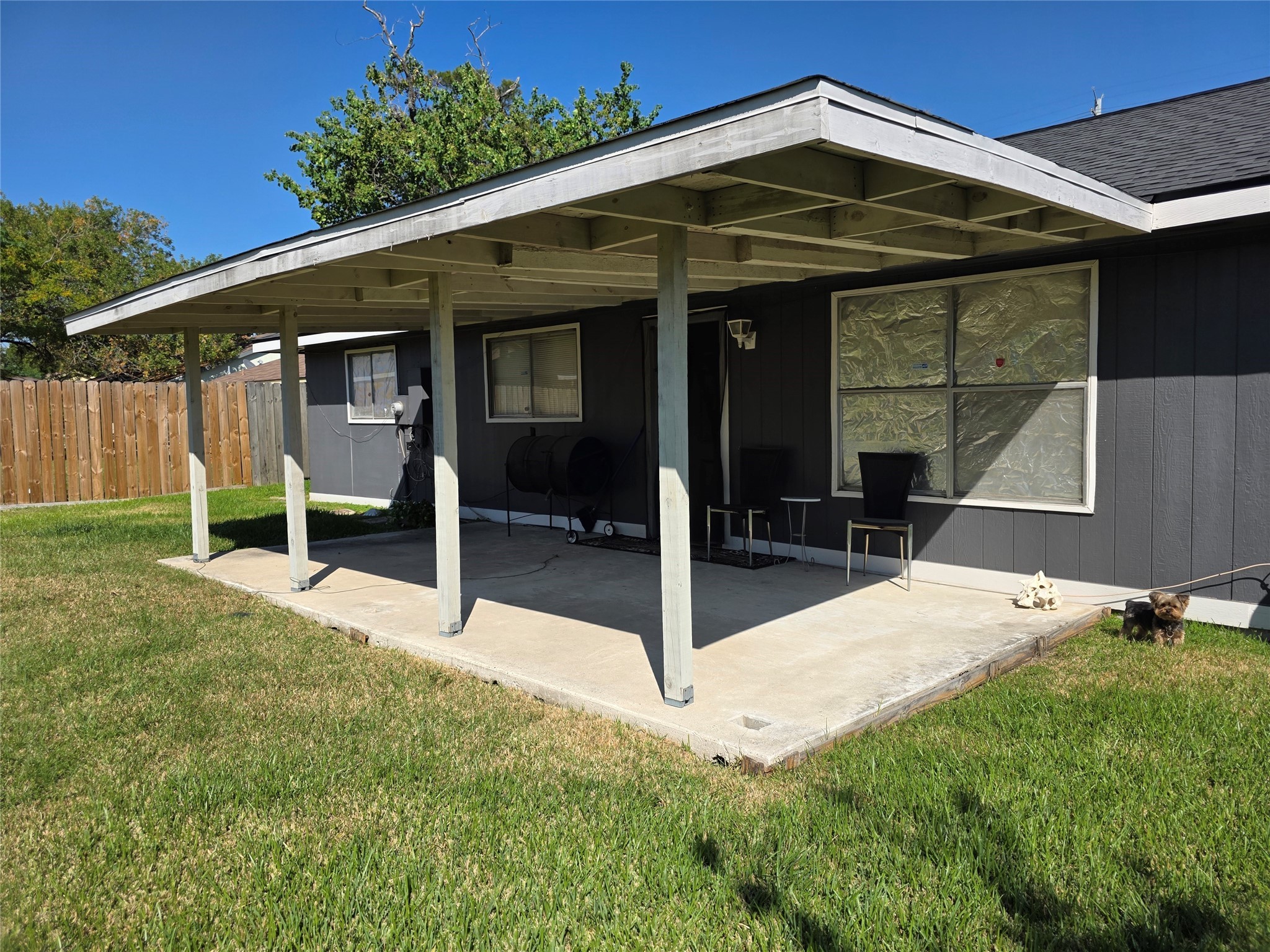 1323 Padstow Lane Channelview, TX 77530 - Photo 11 of 11 This photo shows a backyard with a covered patio attached to a house, featuring a concrete floor and simple patio furniture. The yard is grassy and enclosed by a wooden fence, offering privacy and outdoor space.