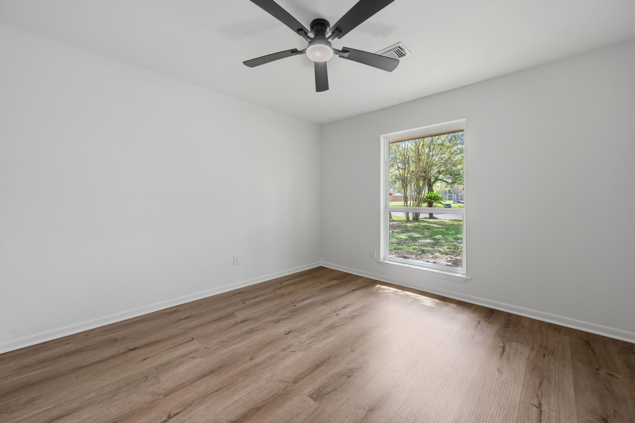 210 Ridgemont Drive Alvin, TX 77511 - Photo 16 of 27 This is a bright, empty room with light-colored walls and wood flooring. It features a ceiling fan and a large window providing a view of a green outdoor space.
