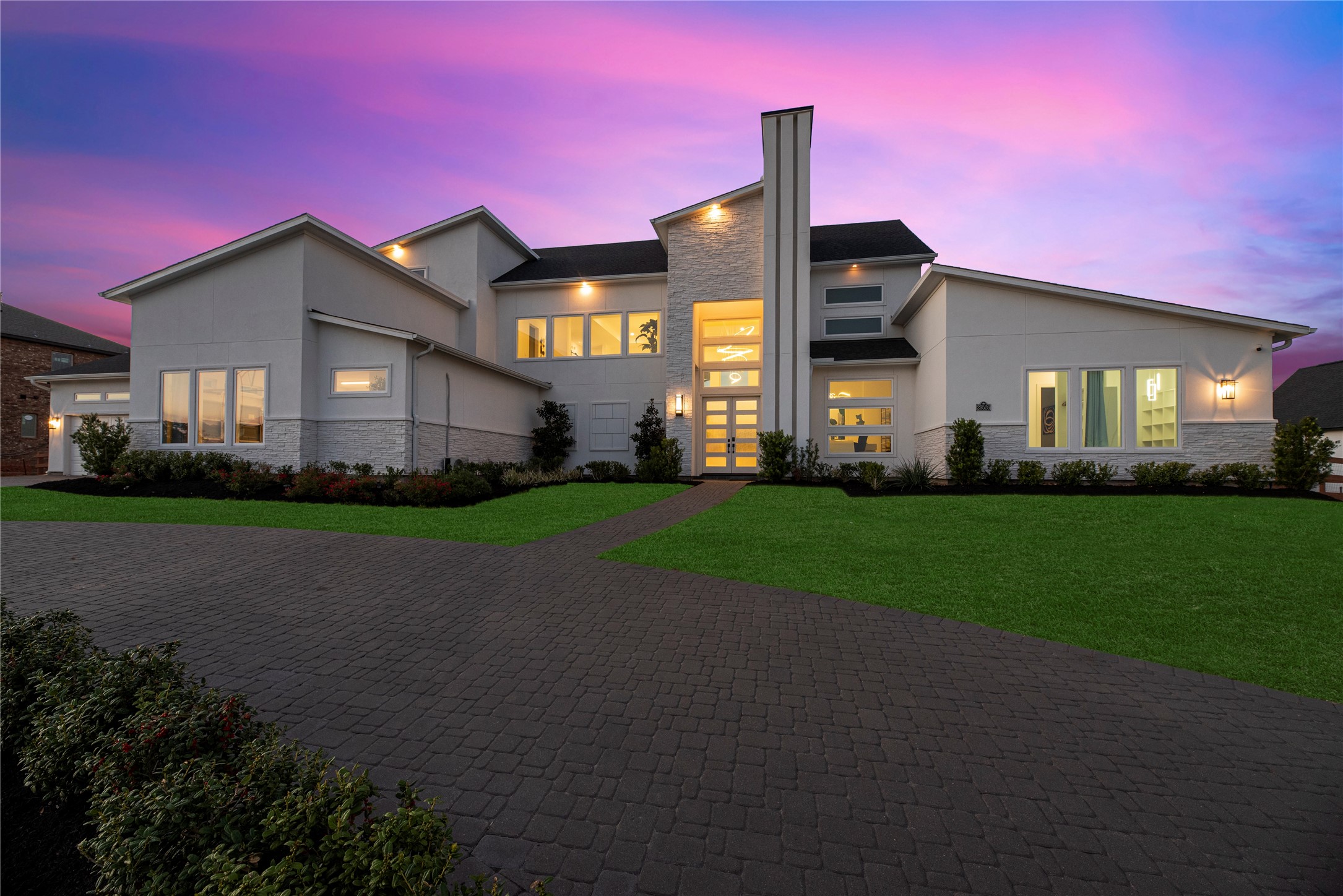 Striking twilight exterior showcasing this modern Fulshear Run home designed by Partners in Building, with clean architectural lines, expansive windows, and a welcoming entry framed by warm lighting. The extended paver driveway and thoughtfully designed elevation create a memorable first impression day or night.