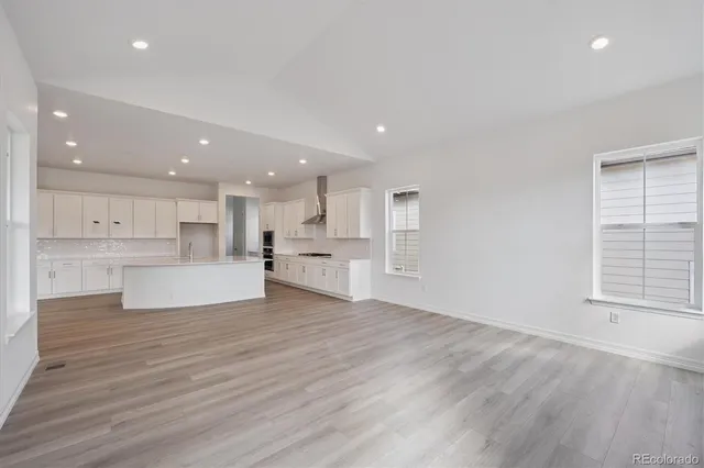 a view of an empty room with wooden floor and kitchen view