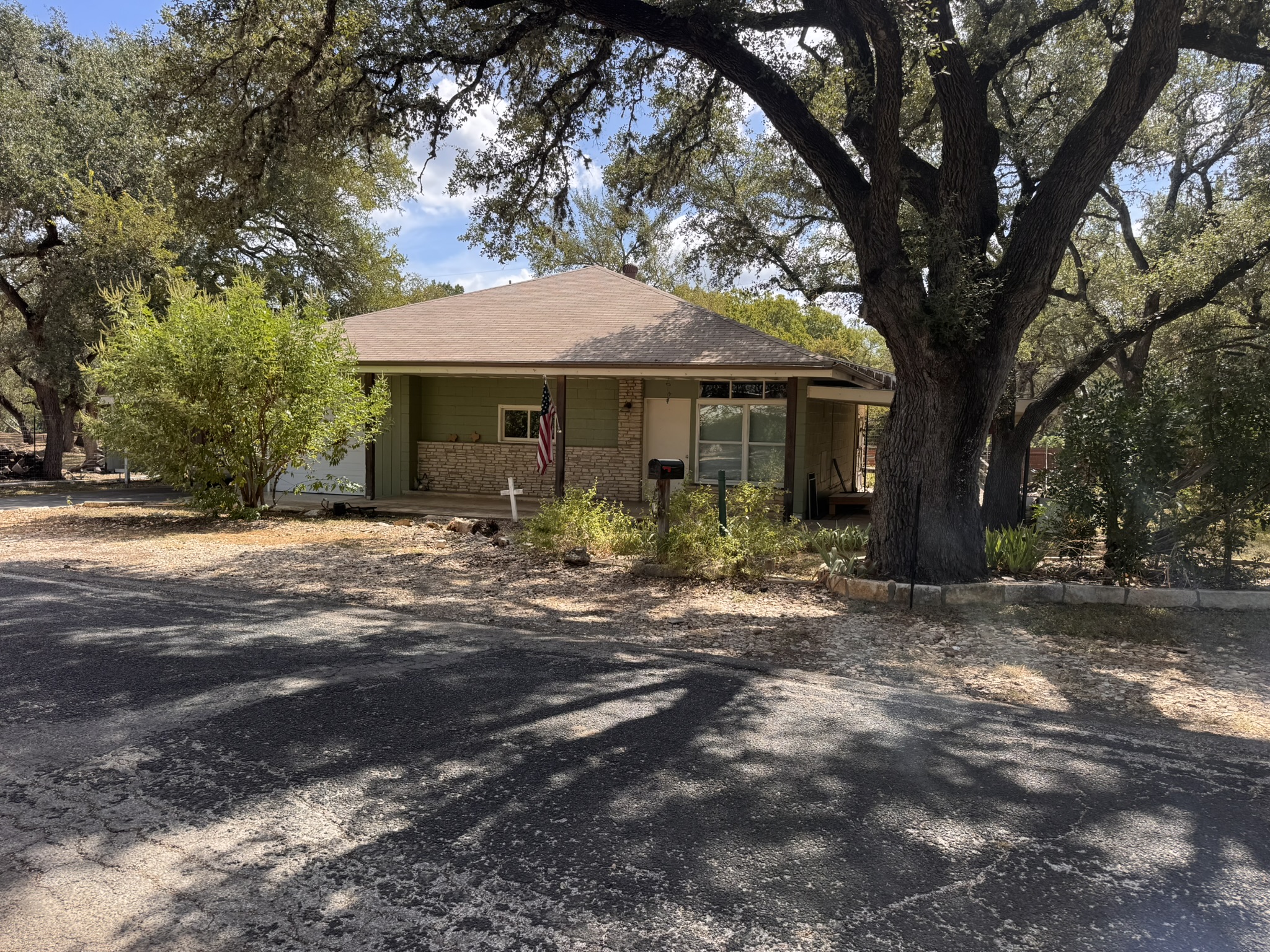 13 Presidio Road Wimberley, TX 78676 - Photo 2 of 7 Mature shade trees frame a classic single-story exterior featuring stone accents and shingle roofing.