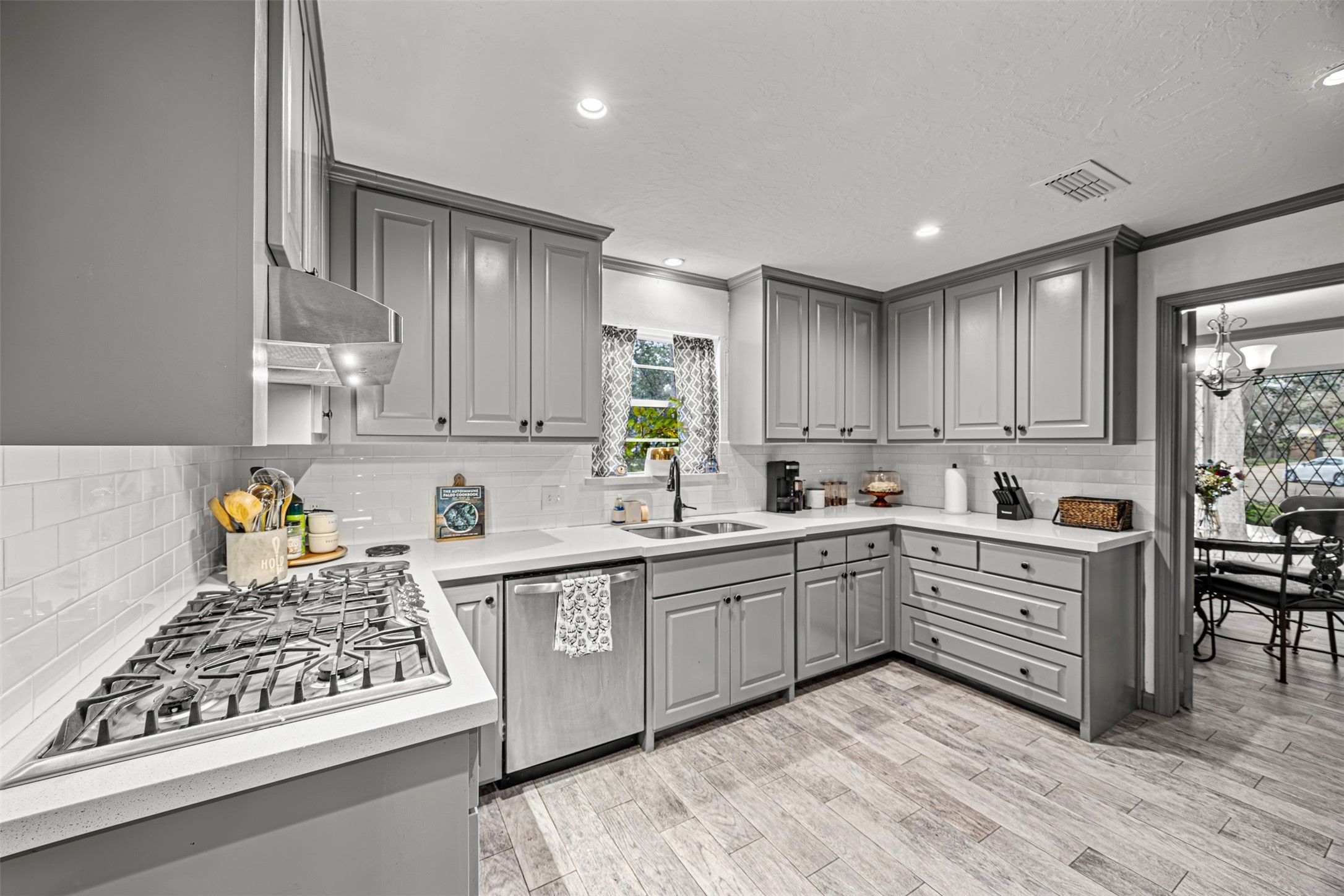 This kitchen features sleek gray cabinetry, a modern gas stove cooktop, and white subway tile backsplash. The spacious layout includes ample counter space and a window above the sink for natural light. Adjacent is a dining area with a decorative chandelier.