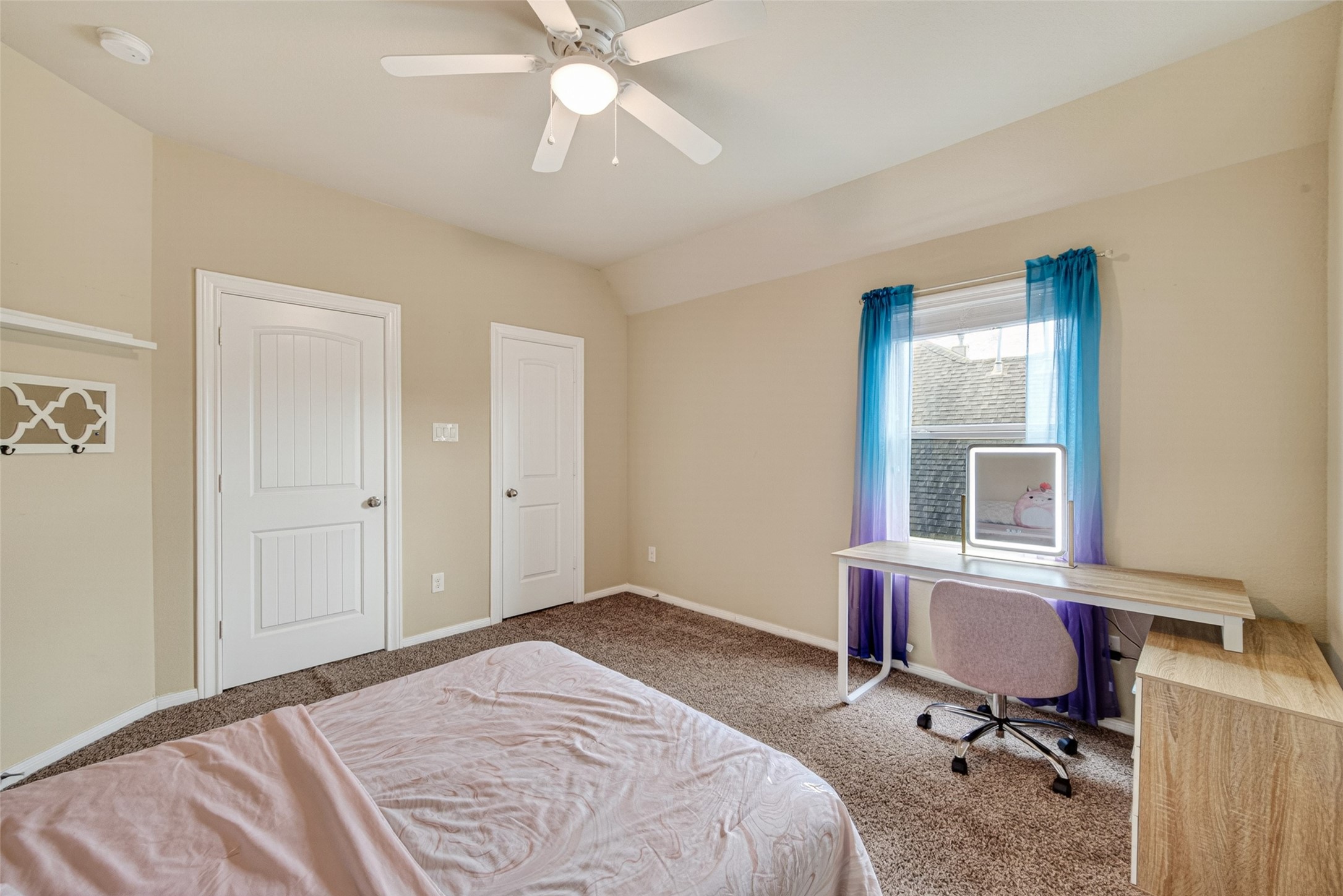 25611 Ellis Ridge Court Katy, TX 77494 - Photo 36 of 49 This bedroom features neutral walls, carpeted flooring, a ceiling fan, and a window with blue curtains. It includes a simple desk and chair, and two white doors, likely for a closet and entry.
