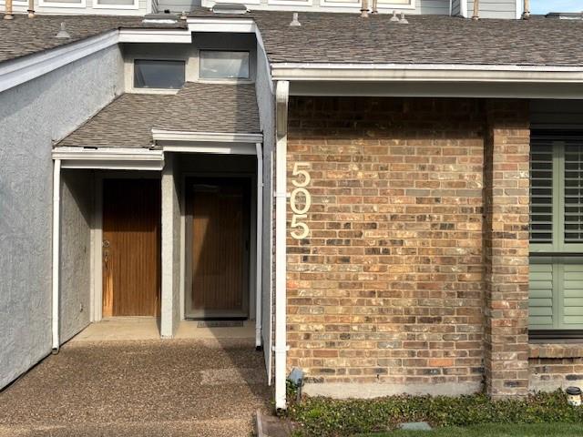 View of exterior entry with brick siding and roof with shingles