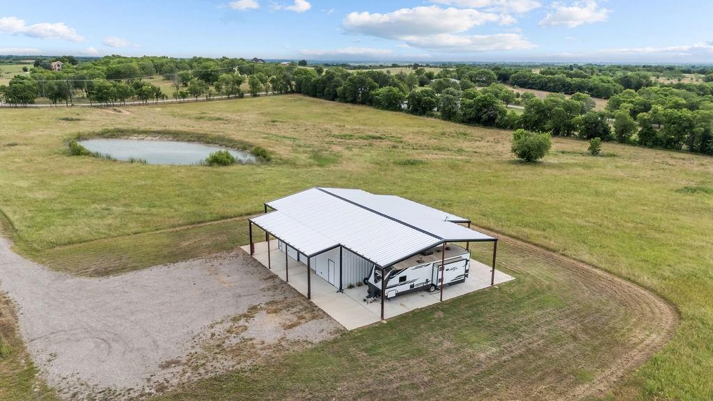 1161 Alsdorf Road Ennis, TX 75119 - Photo 12 of 28 Expansive property featuring a large metal building with an attached covered concrete pad, a gravel driveway, and a pond surrounded by green fields