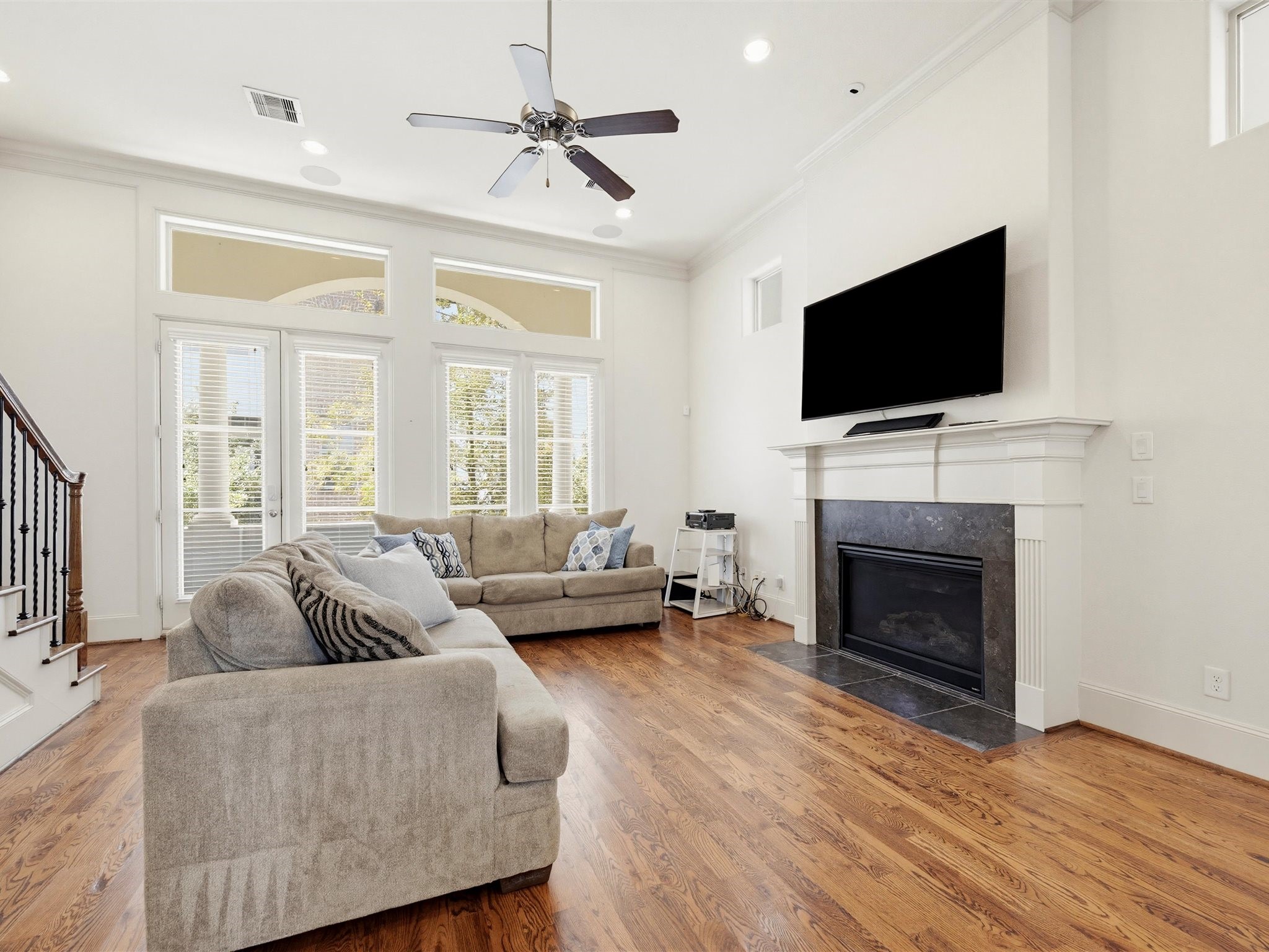 9427 London Bridge Station Houston, TX 77045 - Photo 12 of 30 This living room features hardwood floors, a cozy fireplace with a mounted TV above, and ample natural light from large windows. The space is well-lit with recessed lighting and a ceiling fan, offering a comfortable and inviting atmosphere.