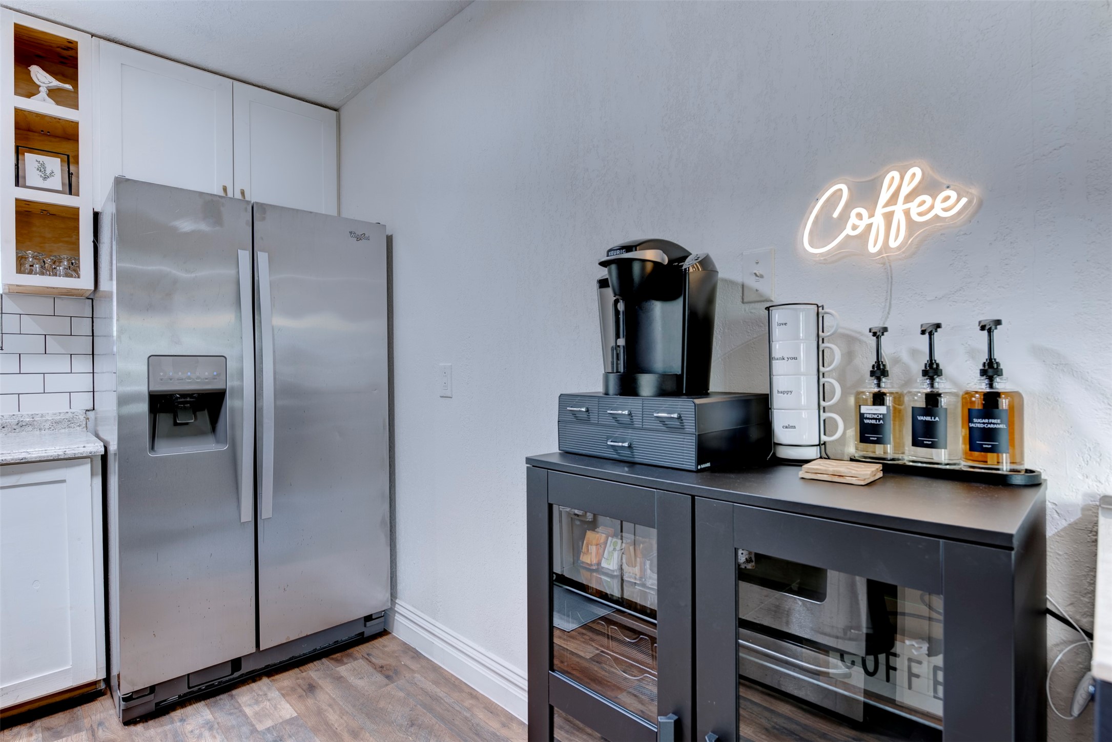 1121 Panama Street Houston, TX 77009 - Photo 13 of 35 Coffee and appliance station close-up showing Whirlpool stainless side-by-side refrigerator with ice and water dispenser, Keurig brewer, labeled syrup dispensers in French Vanilla, Vanilla, and Sugar-Free Salted Caramel, white shaker upper cabinetry with open pine cubby shelving, and white subway tile backsplash.