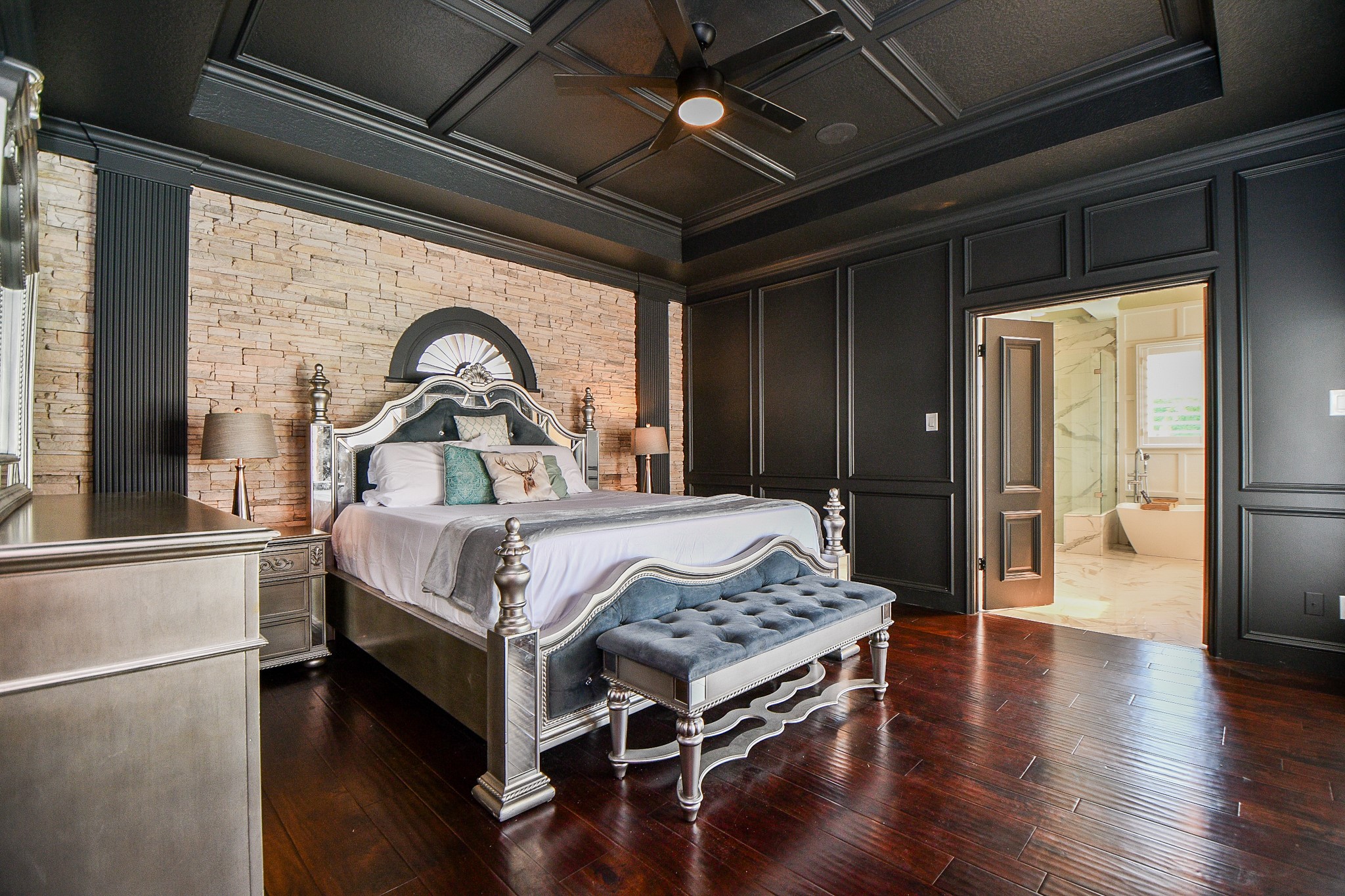 263 Wood Loop Street Houston, TX 77015 - Photo 7 of 45 This elegant master bedroom features dark paneled walls, a coffered ceiling with a ceiling fan, and a stone accent wall. It includes a luxurious bed with a tufted bench, hardwood floors, and an ensuite bathroom with a soaking tub, creating a sophisticated and relaxing retreat.