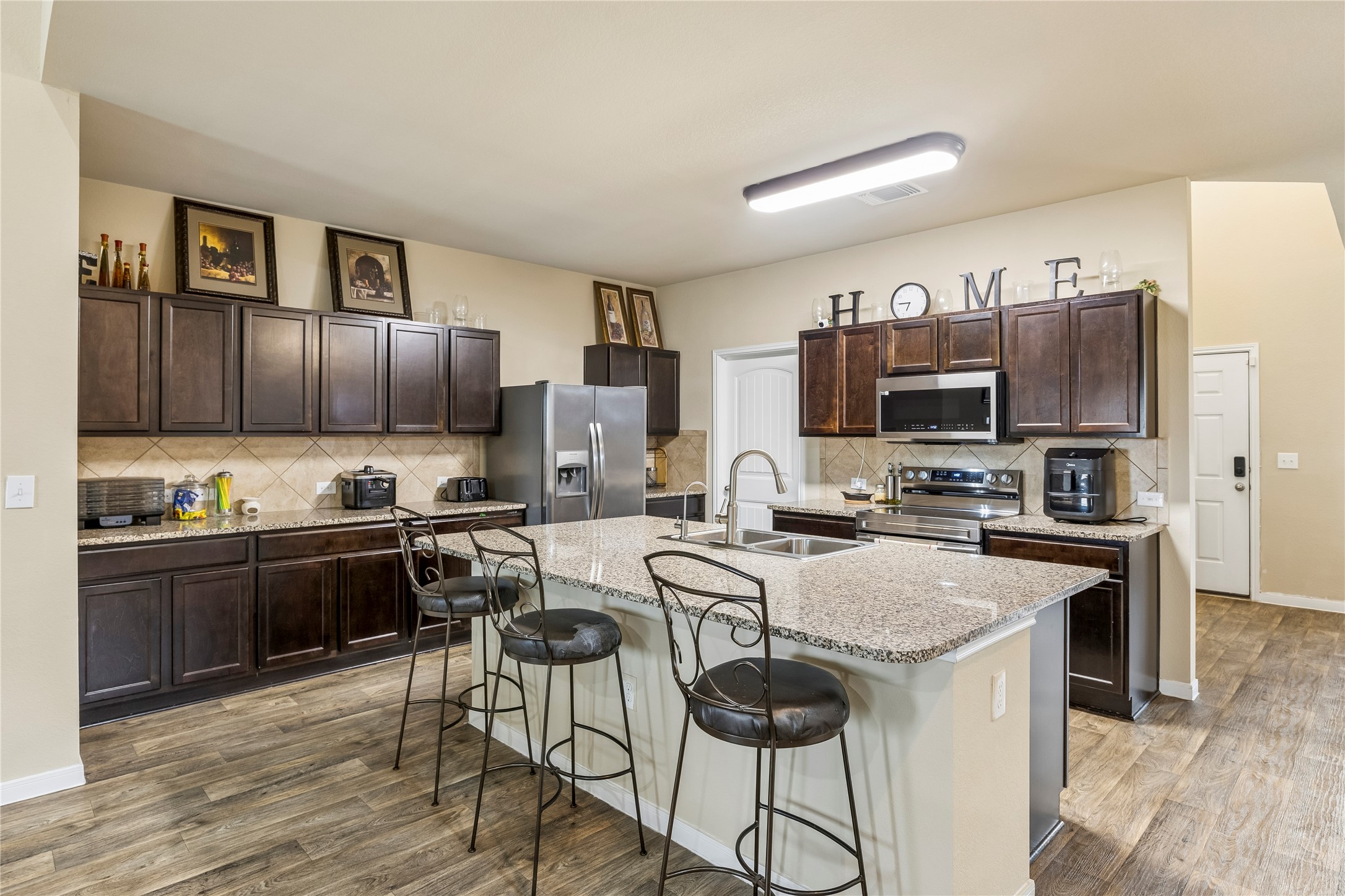 14409 Estuary Road Manor, TX 78653 - Photo 15 of 24 Spacious kitchen featuring an extended center island with integrated double sink, wood-finish flooring, dark wood cabinetry, stainless steel appliances, and granite countertops