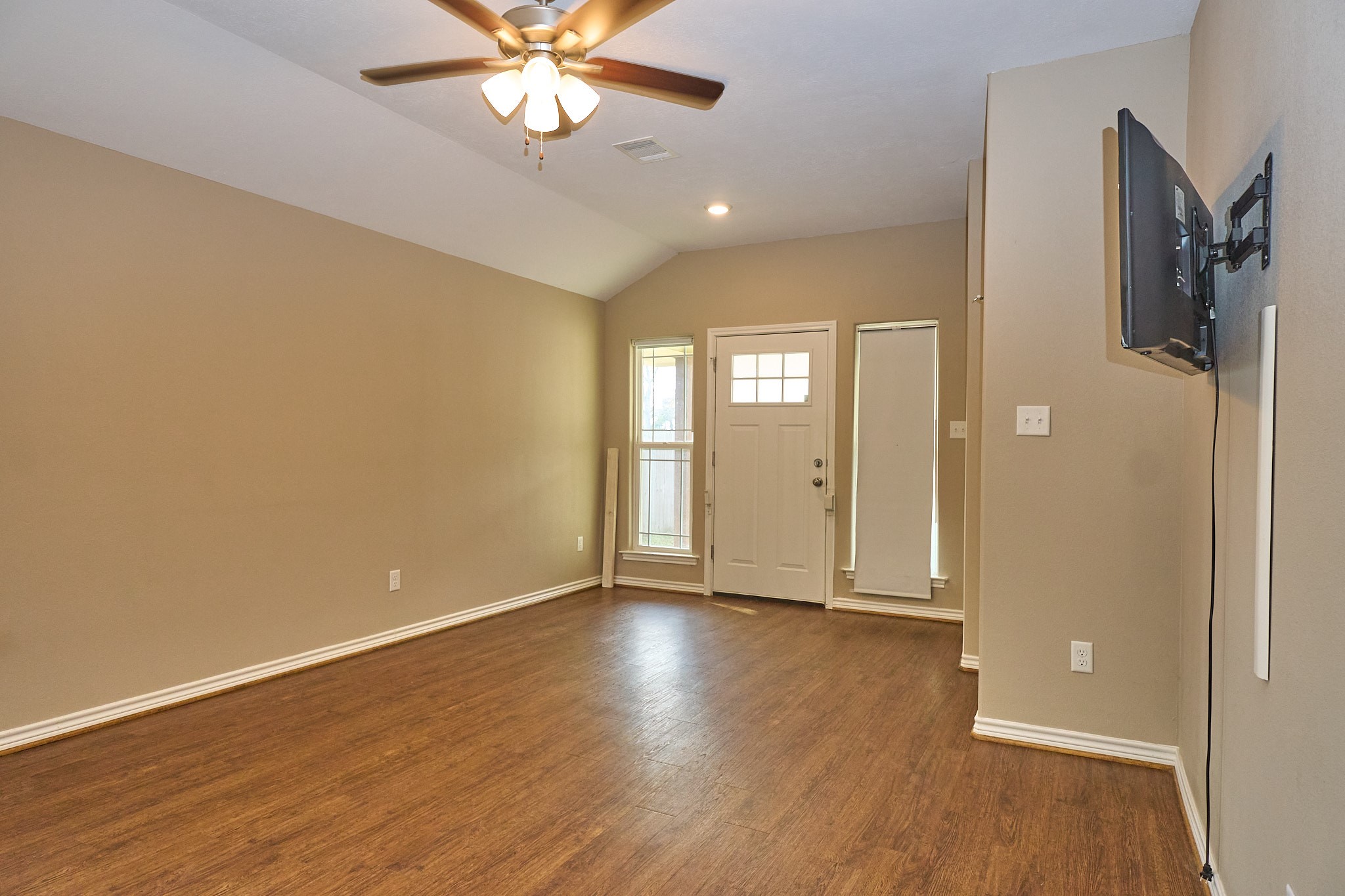 1807 Nuches Lane Bryan, TX 77803 - Photo 6 of 27 This photo showcases a spacious, empty living room with warm wood flooring and neutral walls. It features a ceiling fan, a mounted TV, and ample natural light through a window beside the front door, creating an inviting entryway.