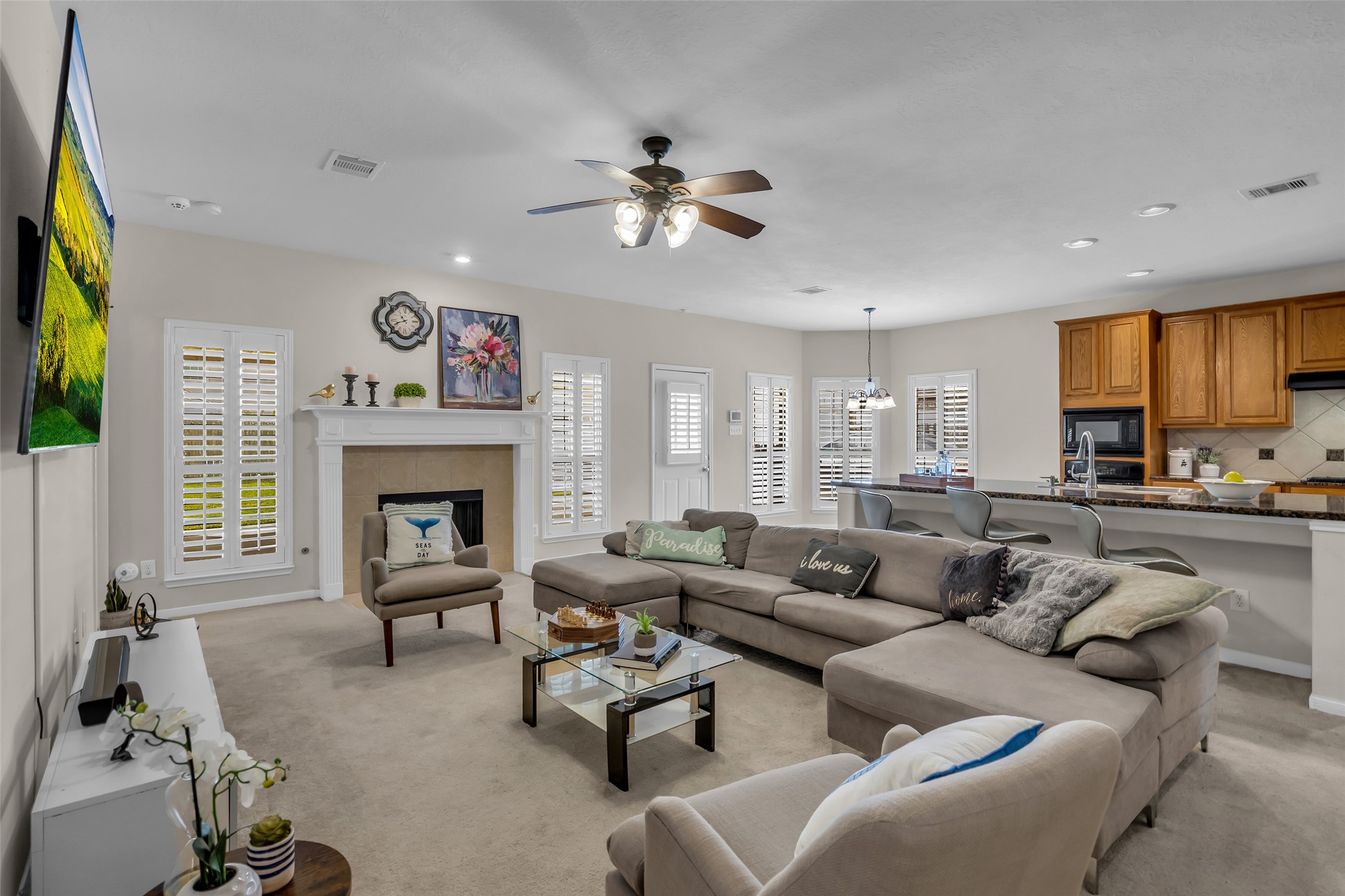 6807 Chapelfield Lane Houston, TX 77049 - Photo 22 of 42 This inviting living space features a cozy fireplace and ample natural light from large windows with plantation shutters. It includes an open-concept layout with a spacious sectional, a modern kitchen with a breakfast bar, and elegant decor, creating a warm and welcoming atmosphere.