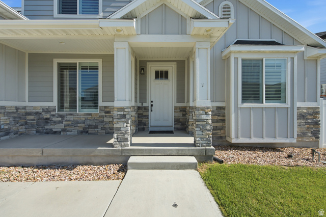 36 West Syracuse Road Vineyard, UT 84059 - Photo 6 of 56 Light gray siding exterior with stone veneer accents, a covered concrete porch, white paneled front door with a transom window, and a bay window featuring louvered blinds