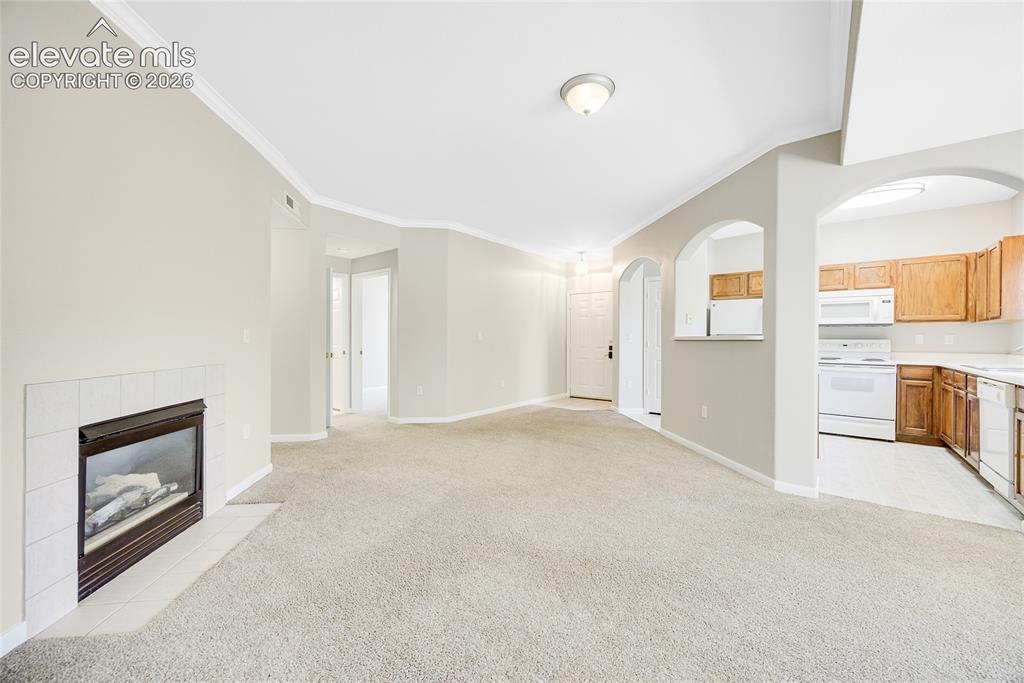 3864 Riviera Grove, Unit 102 Colorado Springs, CO 80922 - Photo 5 of 24 Carpeted living area featuring a fireplace with white tile surround, crown molding, and arched pass-throughs to the kitchen