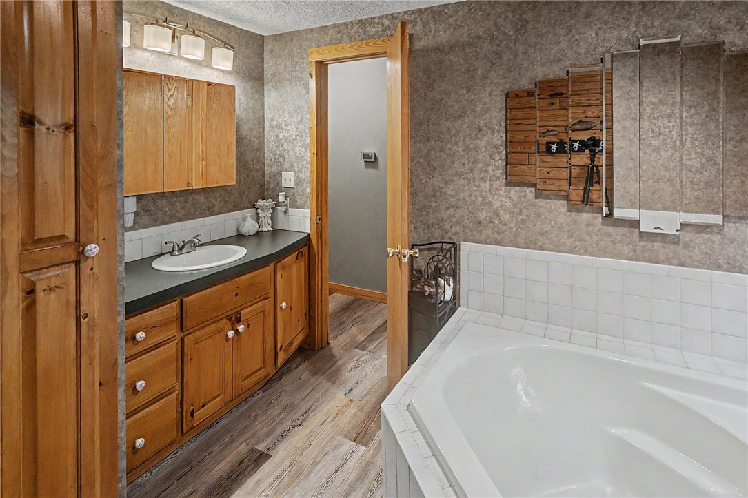 21019 Carter Road West Fork, AR 72774 - Photo 36 of 49 Bathroom featuring a built-in corner soaking tub with white tile surround, a wood-finish vanity with an integrated sink and dark countertop, and wood-finish flooring