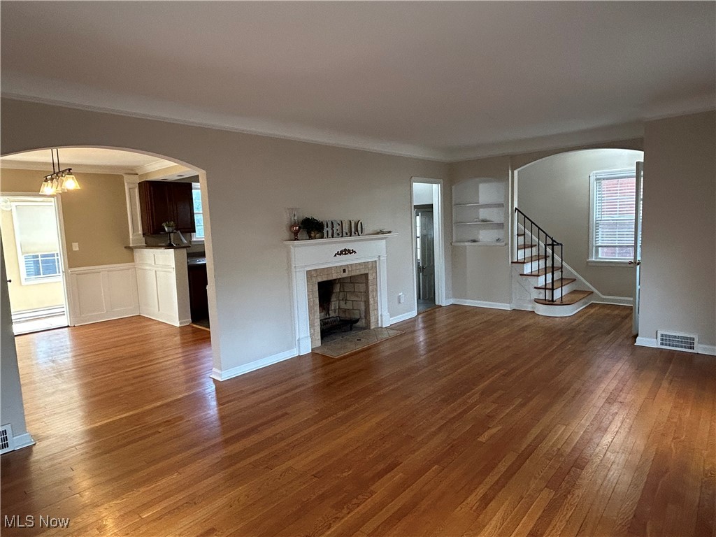 Unfurnished living room with arched walkways, stairway, dark wood-type flooring, a tiled fireplace, and crown molding