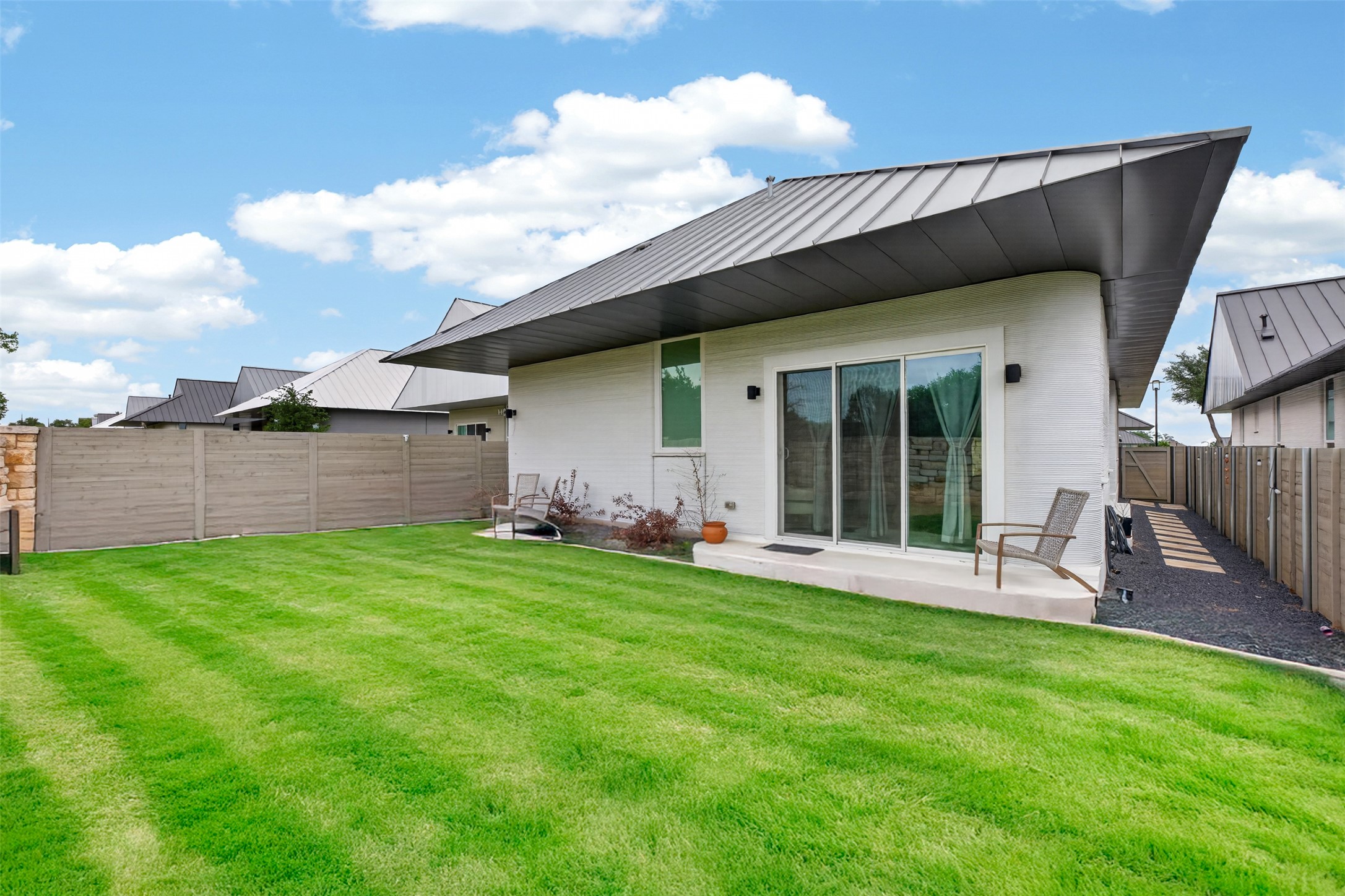 308 Painted Sunset Drive Georgetown, TX 78628 - Photo 23 of 26 Modern exterior with a distinctive dark metal roofline, bright white siding, sliding glass doors to a concrete patio, a spacious green lawn, and a wood-panel fence
